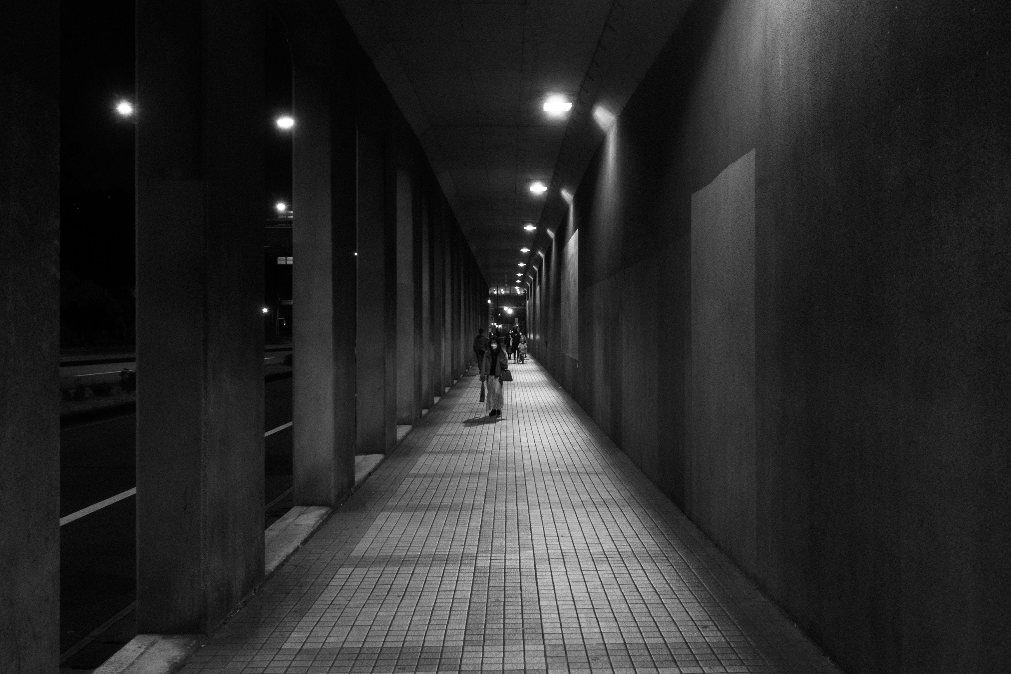 Dimly lit pedestrian tunnel at night with tiled flooring and columned walls. A person walks in the distance, while streetlights illuminate the path. Shadows and light create a pattern on the walls and floor.