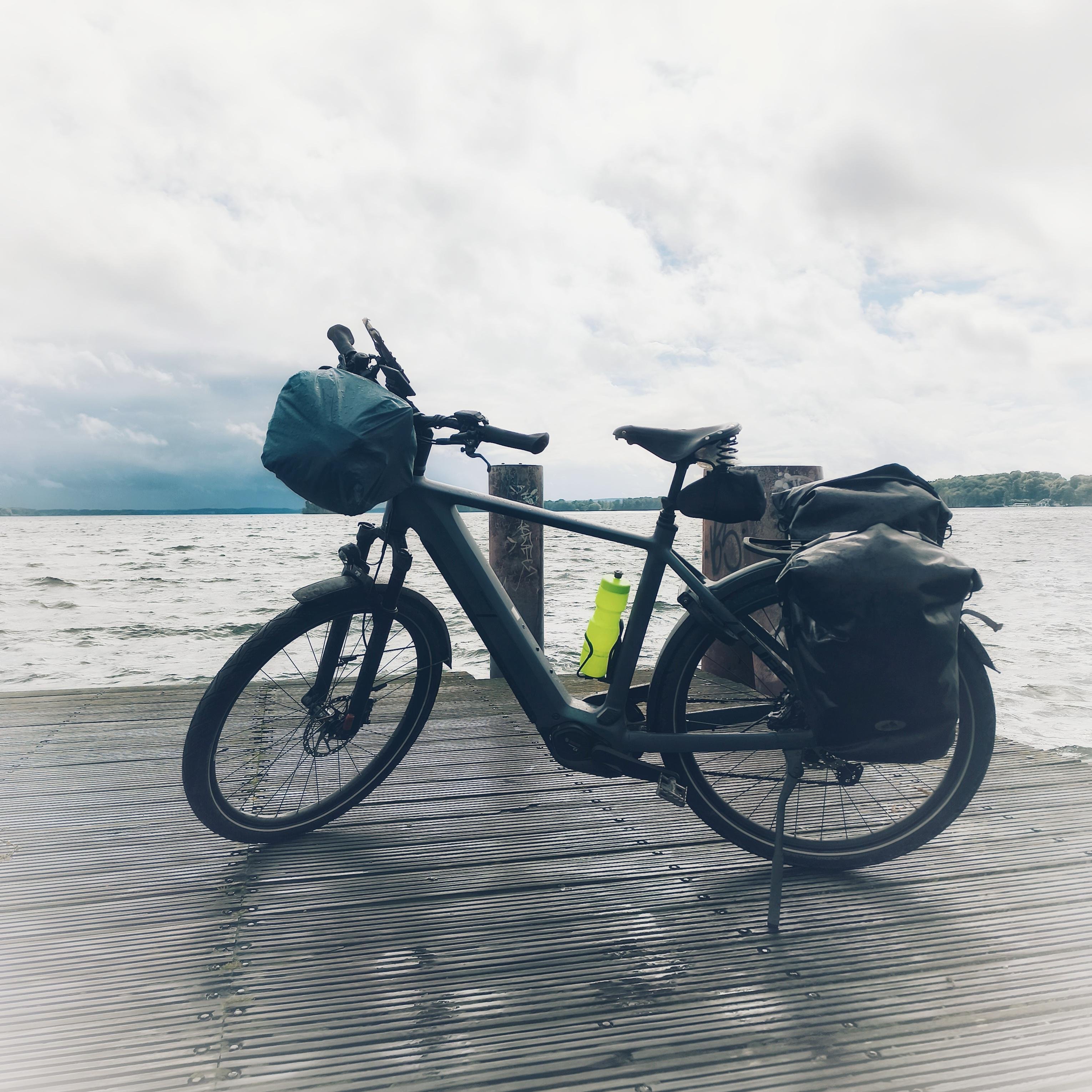 Loaded touring bicycle on a wooden dock by a lake, with panniers on the rear rack, a handlebar bag, and a neon yellow water bottle on the frame; cloudy sky and distant shoreline in the background.
###
Beladenes Tourenrad auf einem Holzsteg an einem See, mit Packtaschen am Gepäckträger, einer Lenkertasche und einer neongelben Trinkflasche am Rahmen; im Hintergrund ein bewölkter Himmel und ein entferntes Ufer.