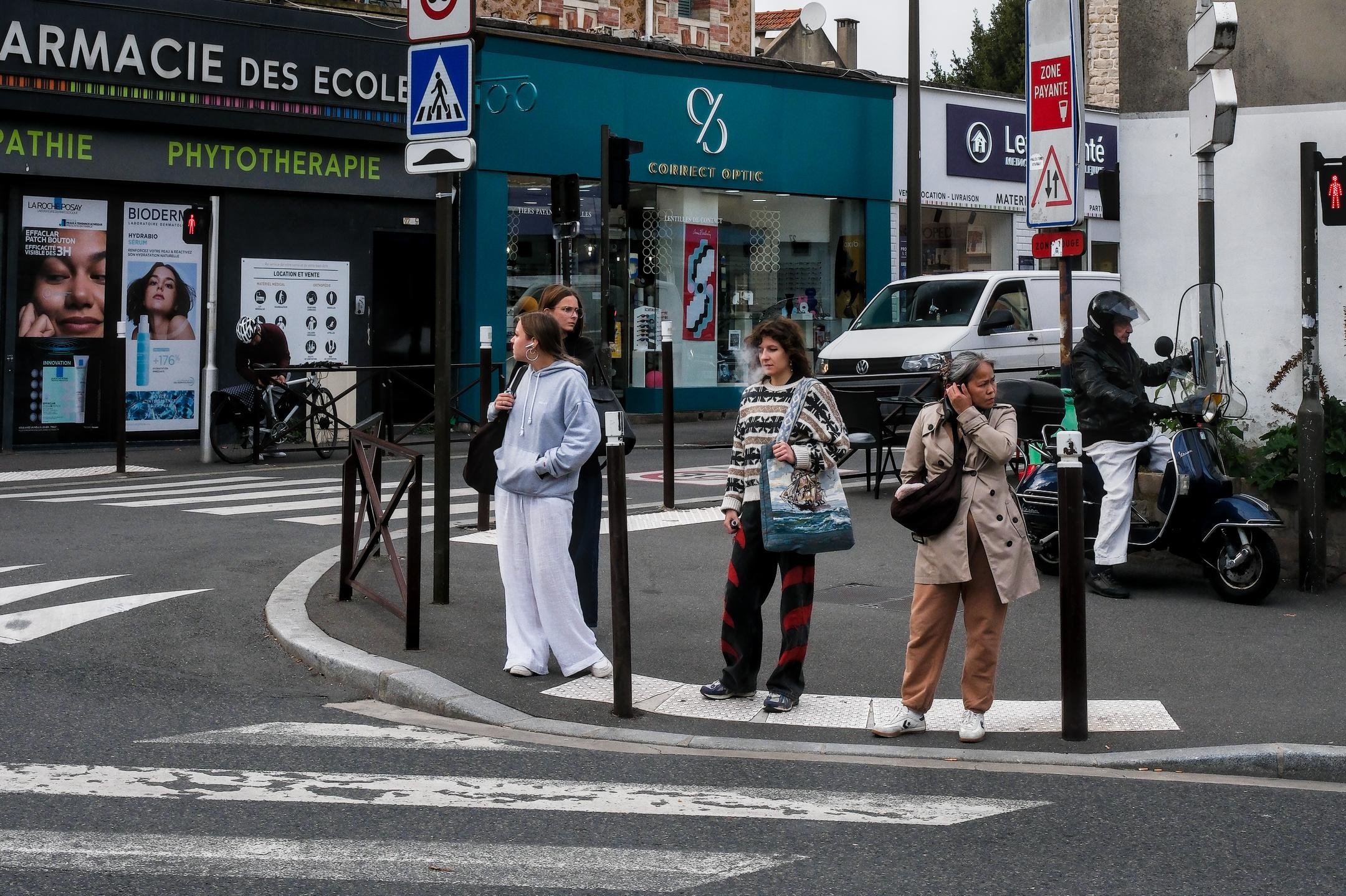 Photo horizontale. 3 femmes attendent à un passage piéton, celle de gauche regarde à gauche, celle de droite, à droite et celle du centre, droit devant elle