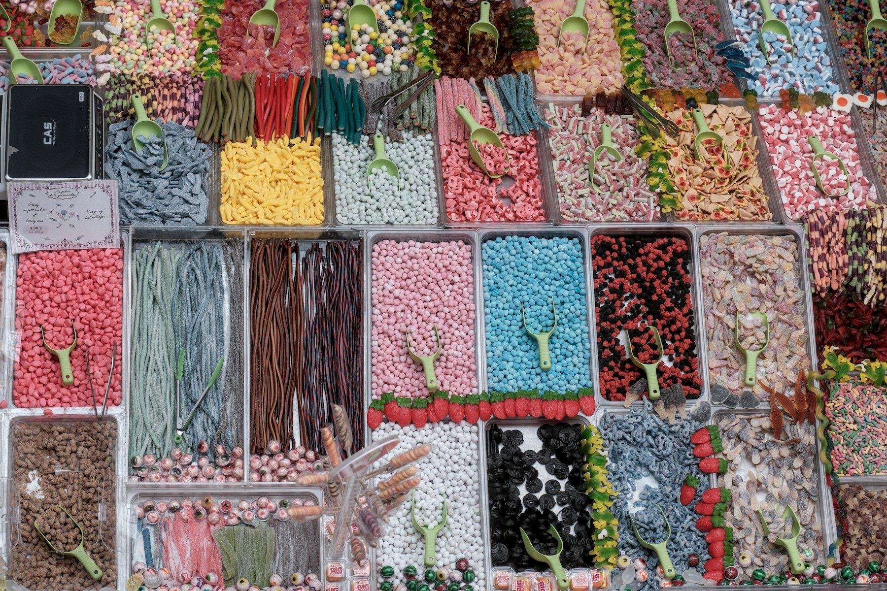 Photo horizontale. Vue de dessus d’un stand de vente de bonbons, plein de bacs, chacun rempli avec un type de sucrerie, faisant une mosaïque de couleurs