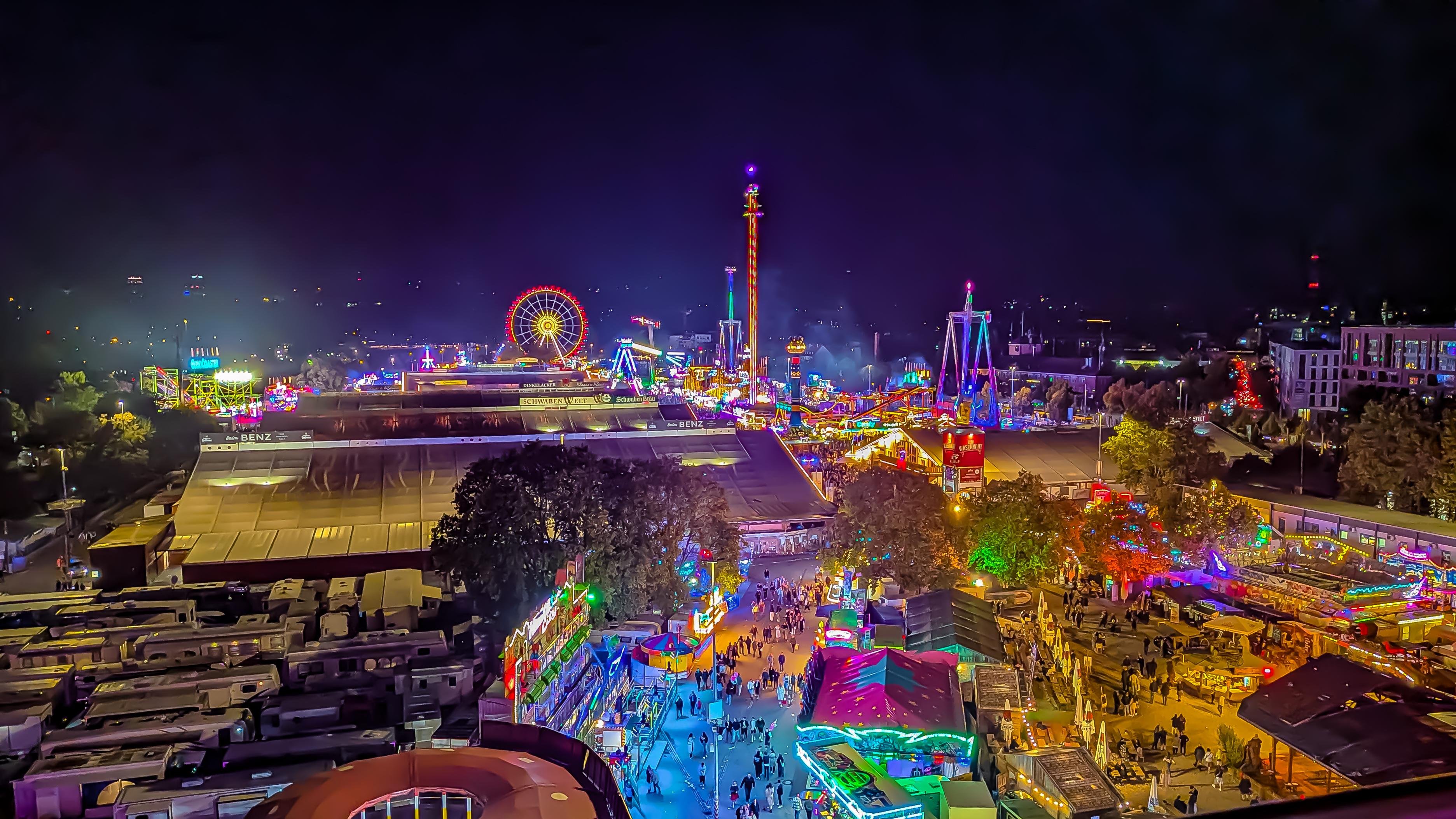 Nighttime amusement park scene with brightly lit rides and attractions. A large, illuminated Ferris wheel and a tall observation tower dominate the skyline. Crowds of people walk through colorful stalls and tents, with games and food vendors visible. Vibrant lights in shades of red, blue, green, and yellow create a lively atmosphere. Trees and buildings are visible in the background under a dark sky.