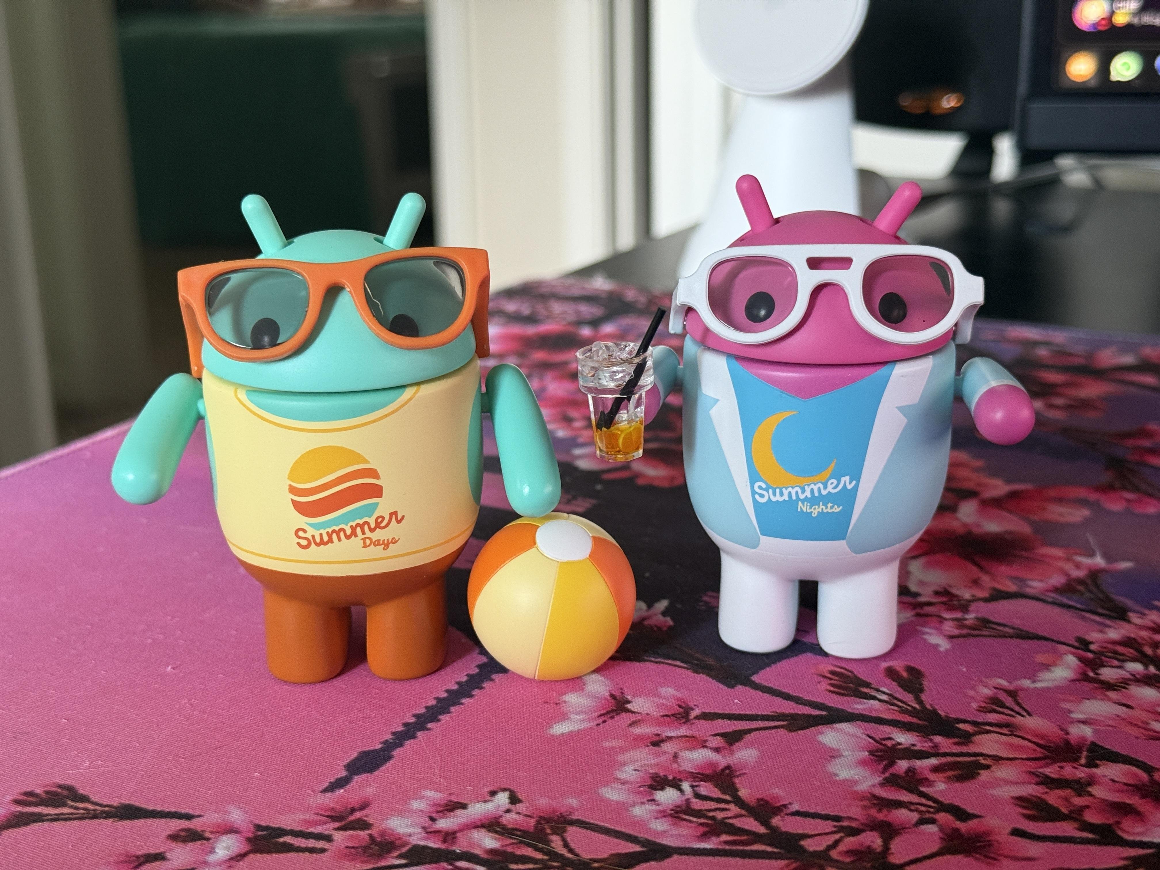 The image features two stylized Android figurines on a pink floral-patterned tablecloth. The figurine on the left is teal with orange sunglasses and a yellow shirt that reads "Summer Days," paired with brown pants. The figurine on the right is pink with white sunglasses and a blue shirt that says "Summer Nights," complemented by a white jacket. Both have antennae on their heads. A small beach ball with orange, yellow, and white stripes is placed between them. The figurine on the right holds a small glass with a straw, suggesting a drink. The background includes a white fan and a computer monitor, adding context to the setting.