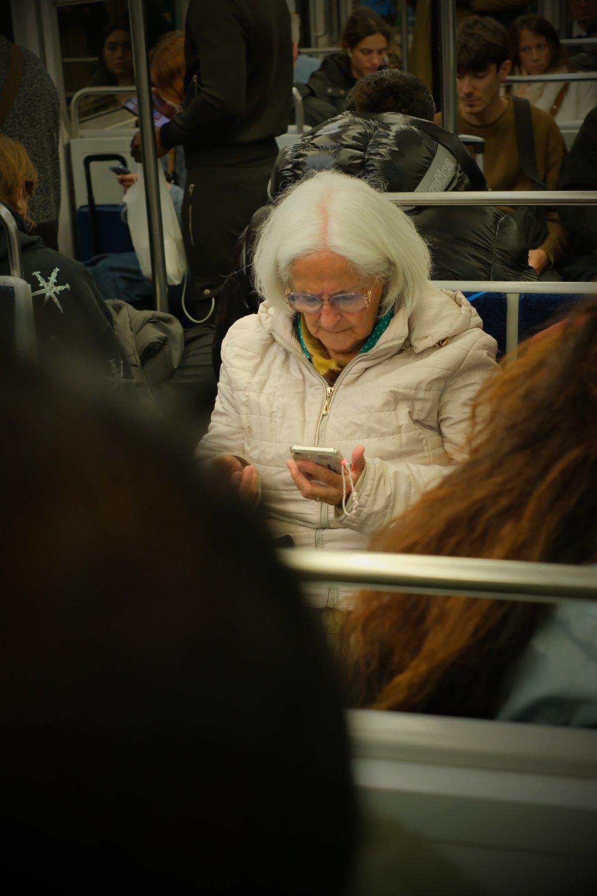 Photo verticale. Une dame au cheveux blanc, manteau blanc, regarde son téléphone, qui a une petite chaînette en petites perles blanches