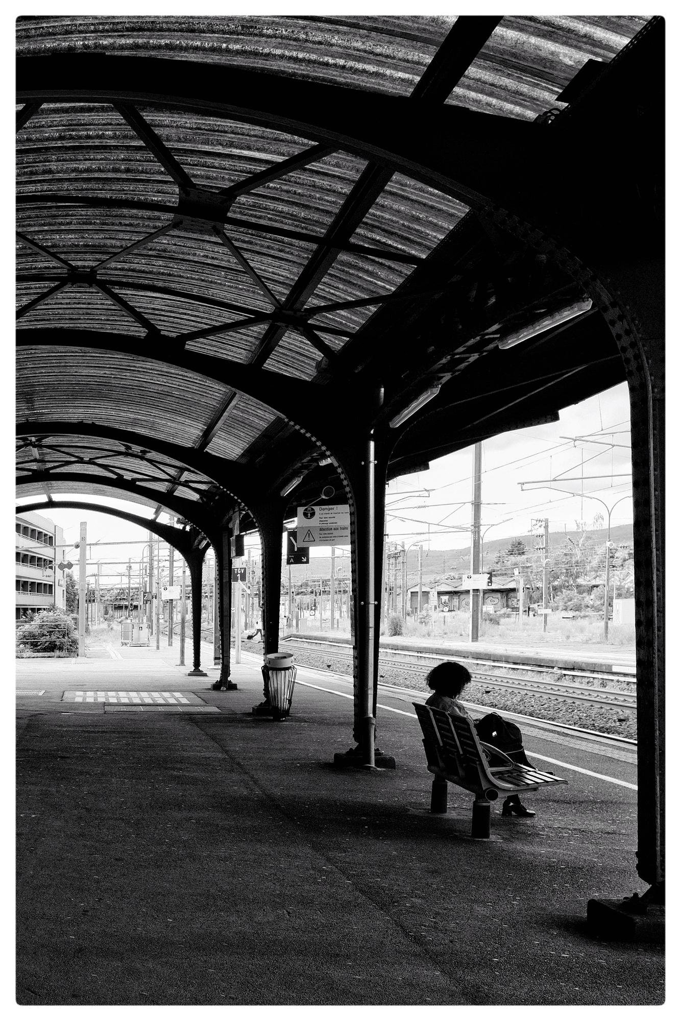 Black and white photo of a train station platform with an arched metal roof. A person sits on a bench to the right, reading. Train tracks run parallel to the platform. Signs and a trash bin are visible under the roof. Buildings and poles are in the background.