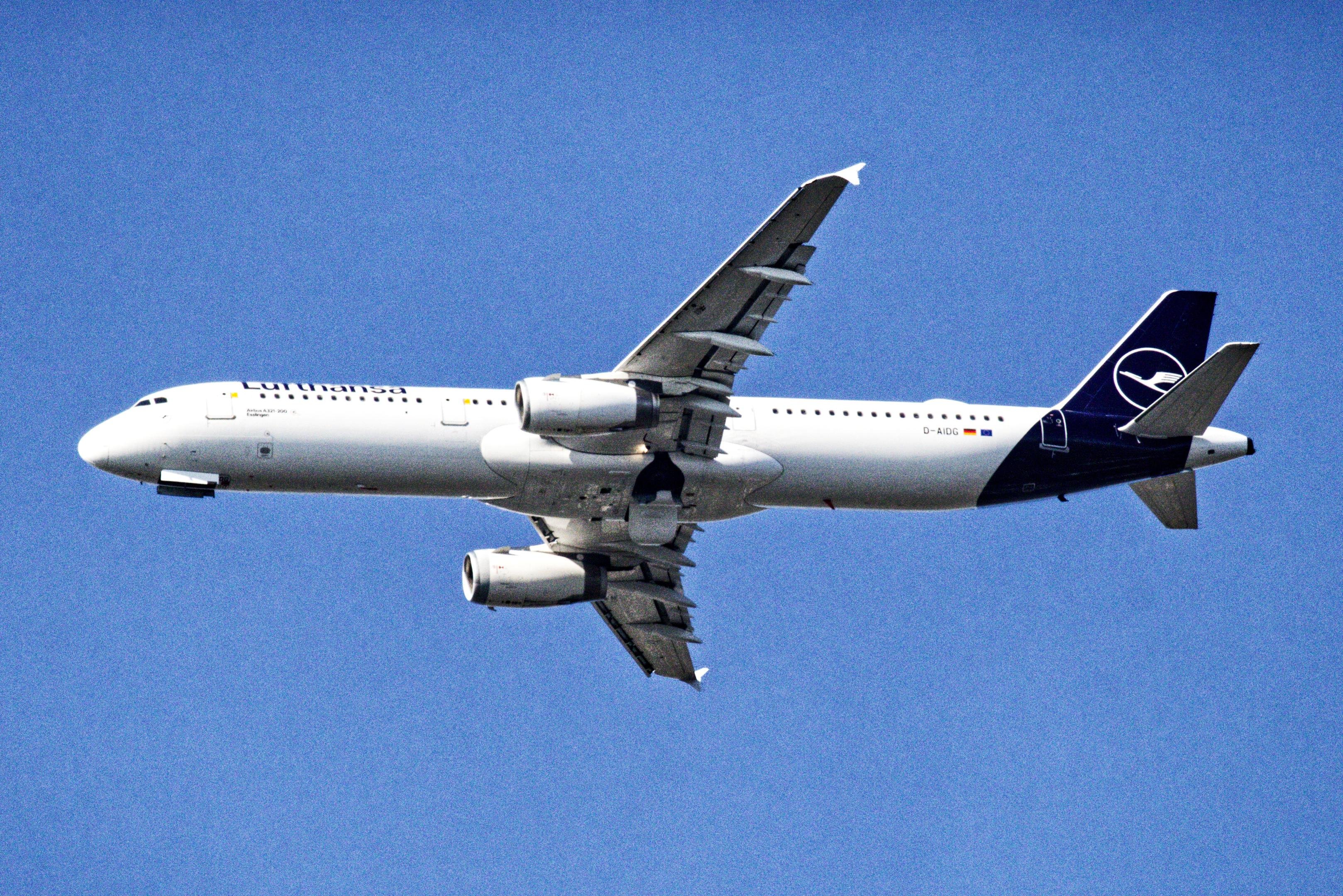 Side view of a Lufthansa passenger jet in flight against a clear blue sky, with landing gear extended and a dark blue tail fin bearing the airline logo.