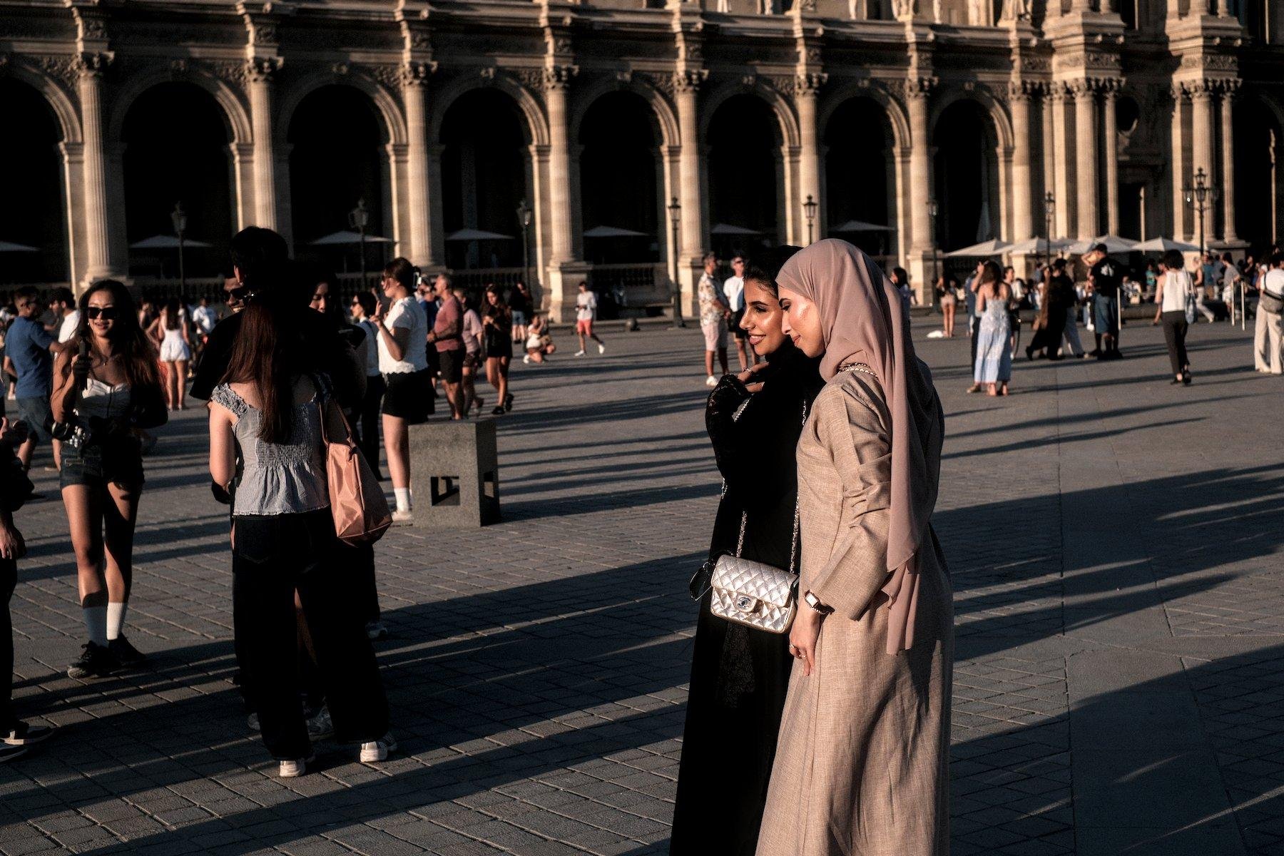 Horizontal photo. 2 women posing together on the Louvre’s place, with many people walking around, minding their own business, on a dawn sunlight