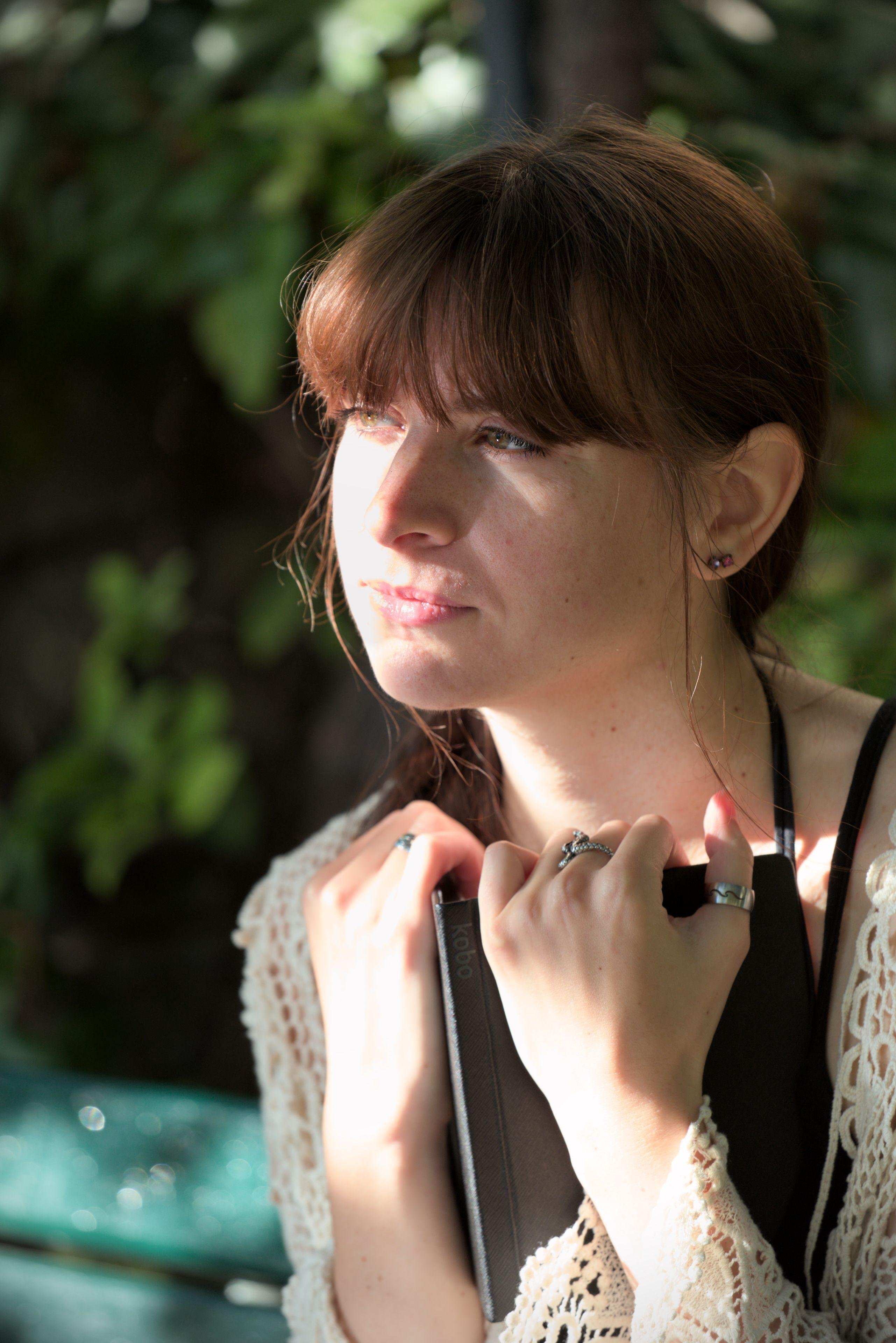Photo format portrait d’une jeune femme brune aux cheveux longs coiffés en une natte qui tombe devant son épaule droite. Elle est habillée d’un débardeur noir à fines bretelles et un gilet fait en crochet avec du fil blanc au motif fleuri. Elle tiens sa liseuse électronique contre son torse des deux mains et regarde au loin, comme méditant sur ce qu’elle vient de lire