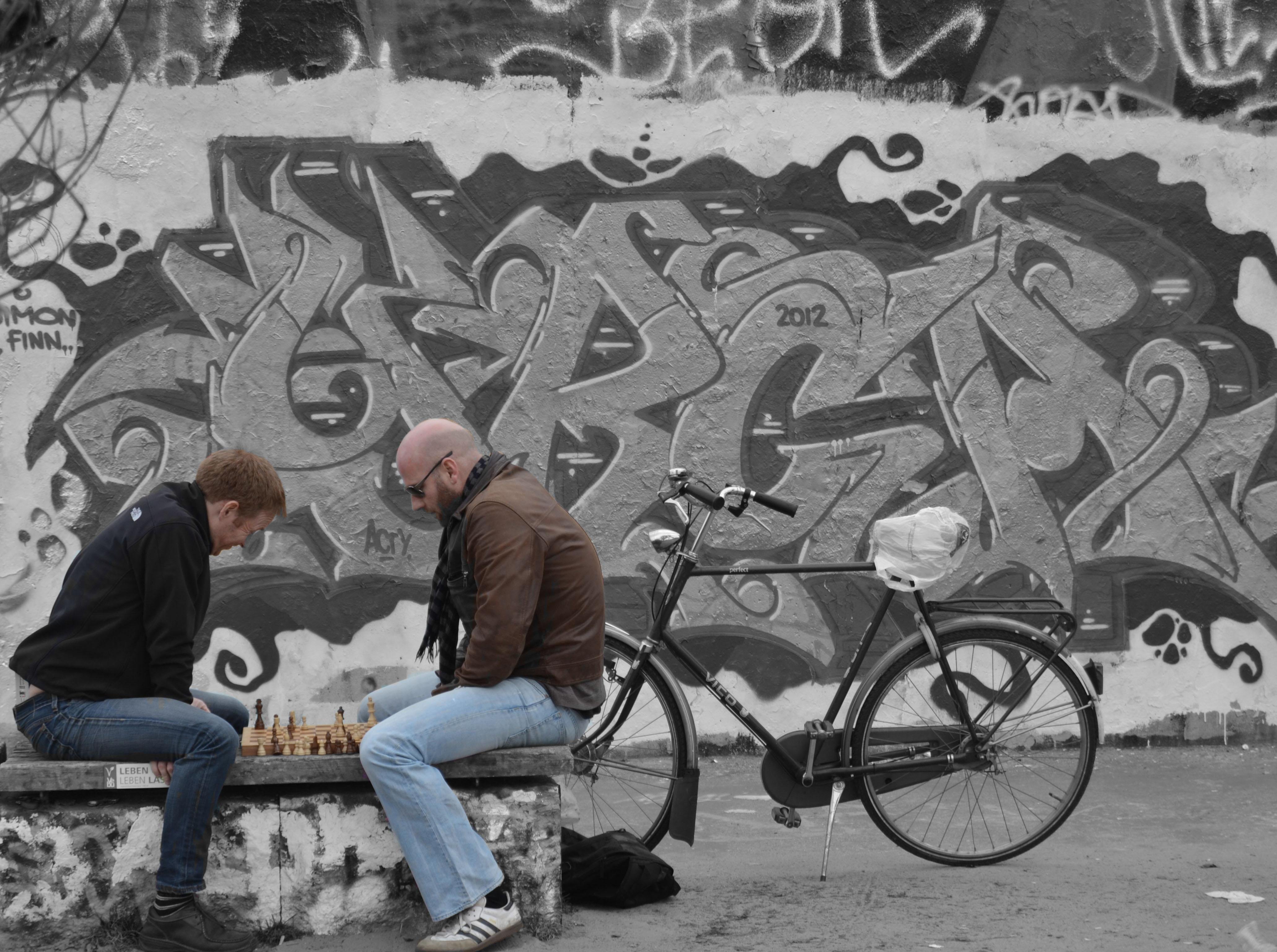 The image depicts two people sitting on a bench playing chess. Behind them, there is a wall with graffiti art. Next to the bench, there is a bicycle. The background seems to be in black and white, while the people, the chessboard, and the bicycle are in color.