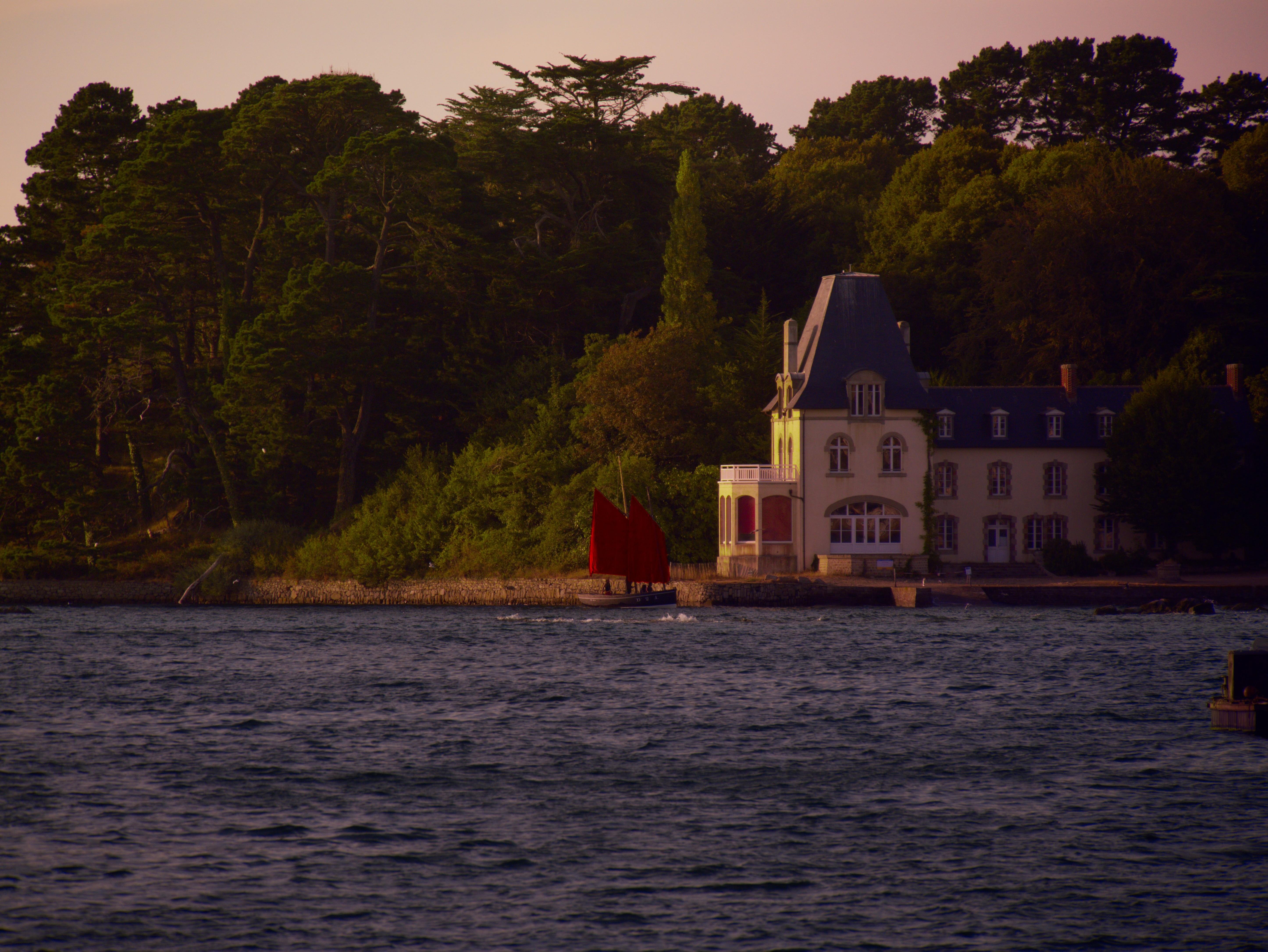 Stone waterfront mansion with a steep dark roof at the edge of a shoreline, viewed across rippling water; a small sailboat with bright red sails passes in front under a pale sky.