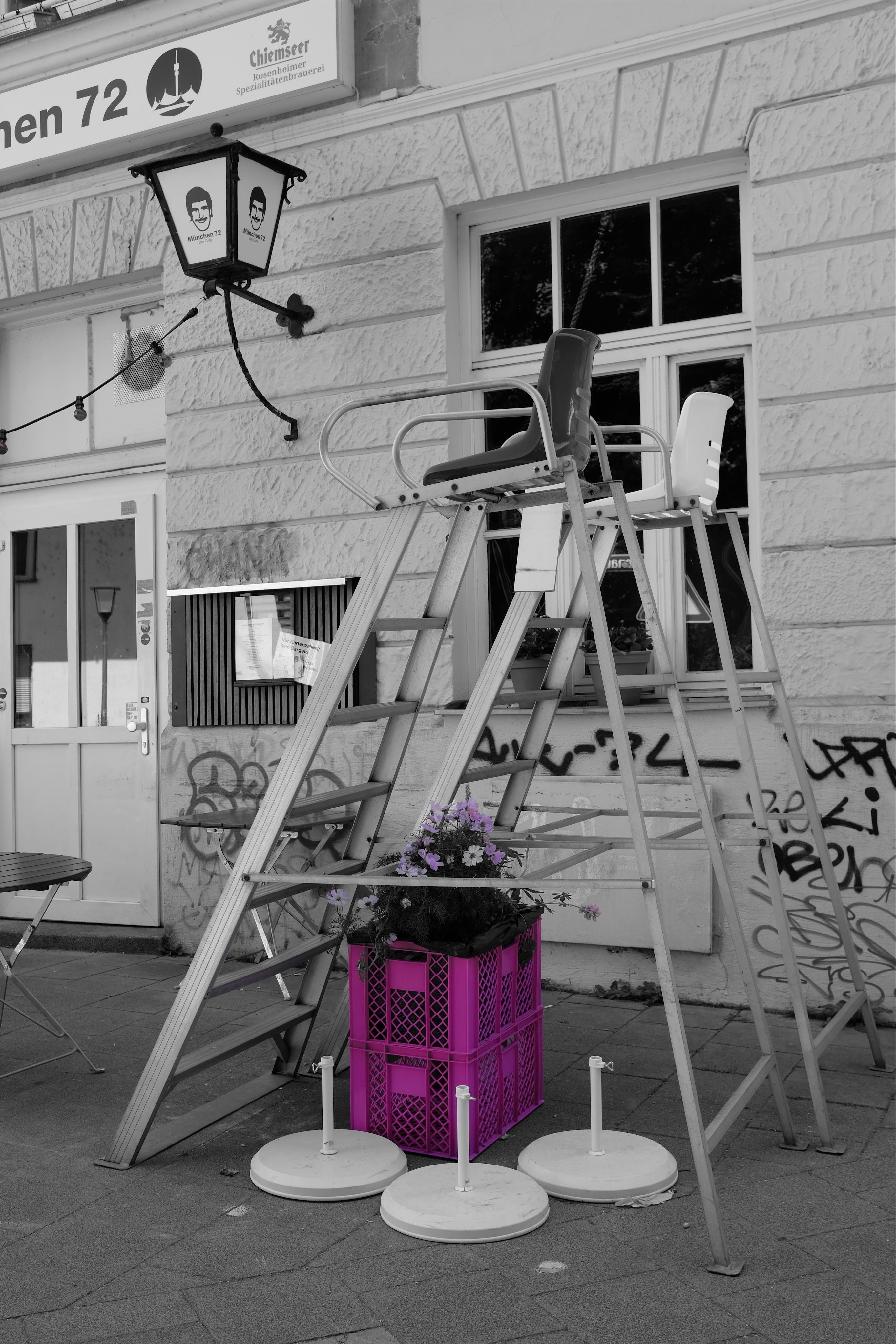 Two metal lifeguard-style chairs, each with a ladder. Both are standing on a sidewalk in front of a graffiti-marked facade of a restaurant/bar. Beneath the chair in the foreground, a purple crate holding flowers sits under the chair, with white umbrella bases on the ground nearby.
Due to partial filtering for purple hues within the camera, only the create and the flowers kept their purple/pink colour, while all the rest of the image is black and white.