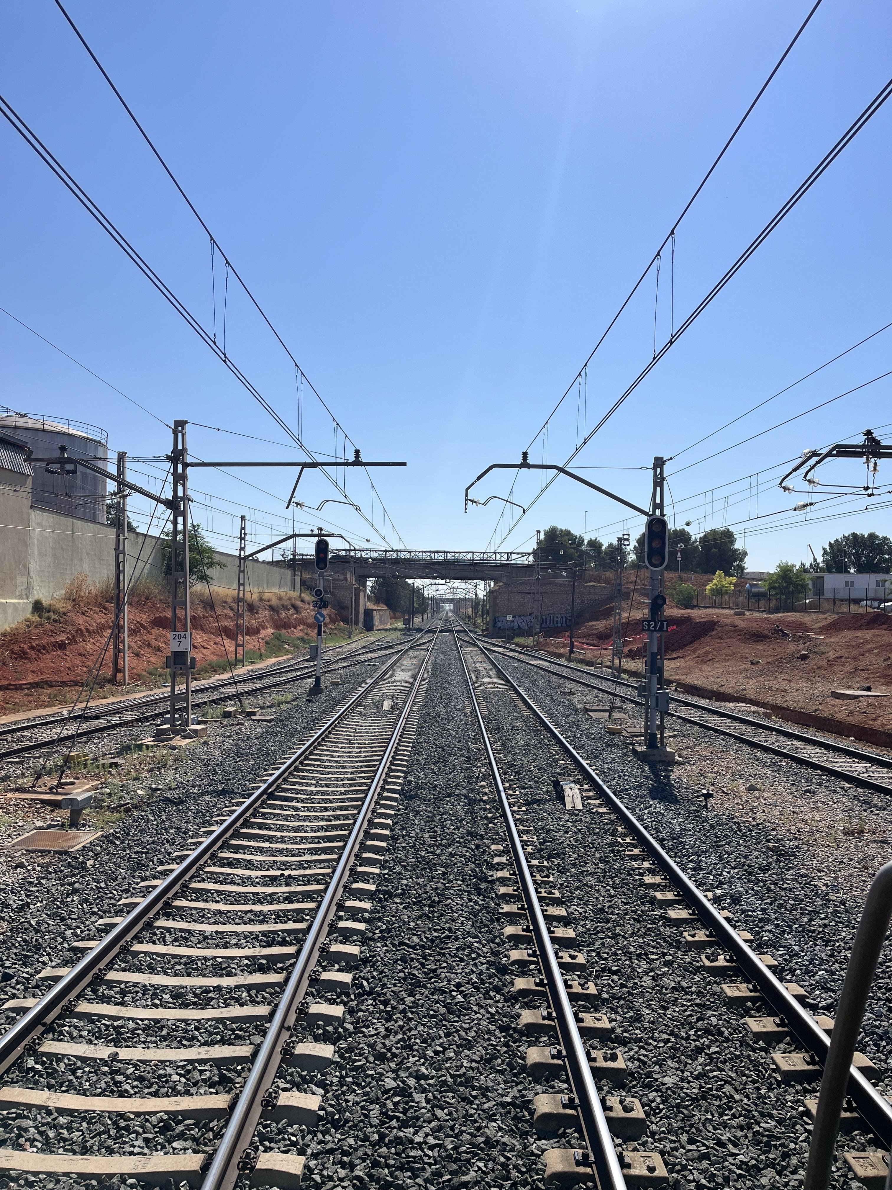 Railway tracks stretching into the distance under a clear blue sky. Overhead power lines and support structures span across the tracks. Multiple sets of tracks are visible, flanked by rough gravel and sparse vegetation. A bridge crosses over the tracks in the distance, and signals are positioned alongside the tracks.