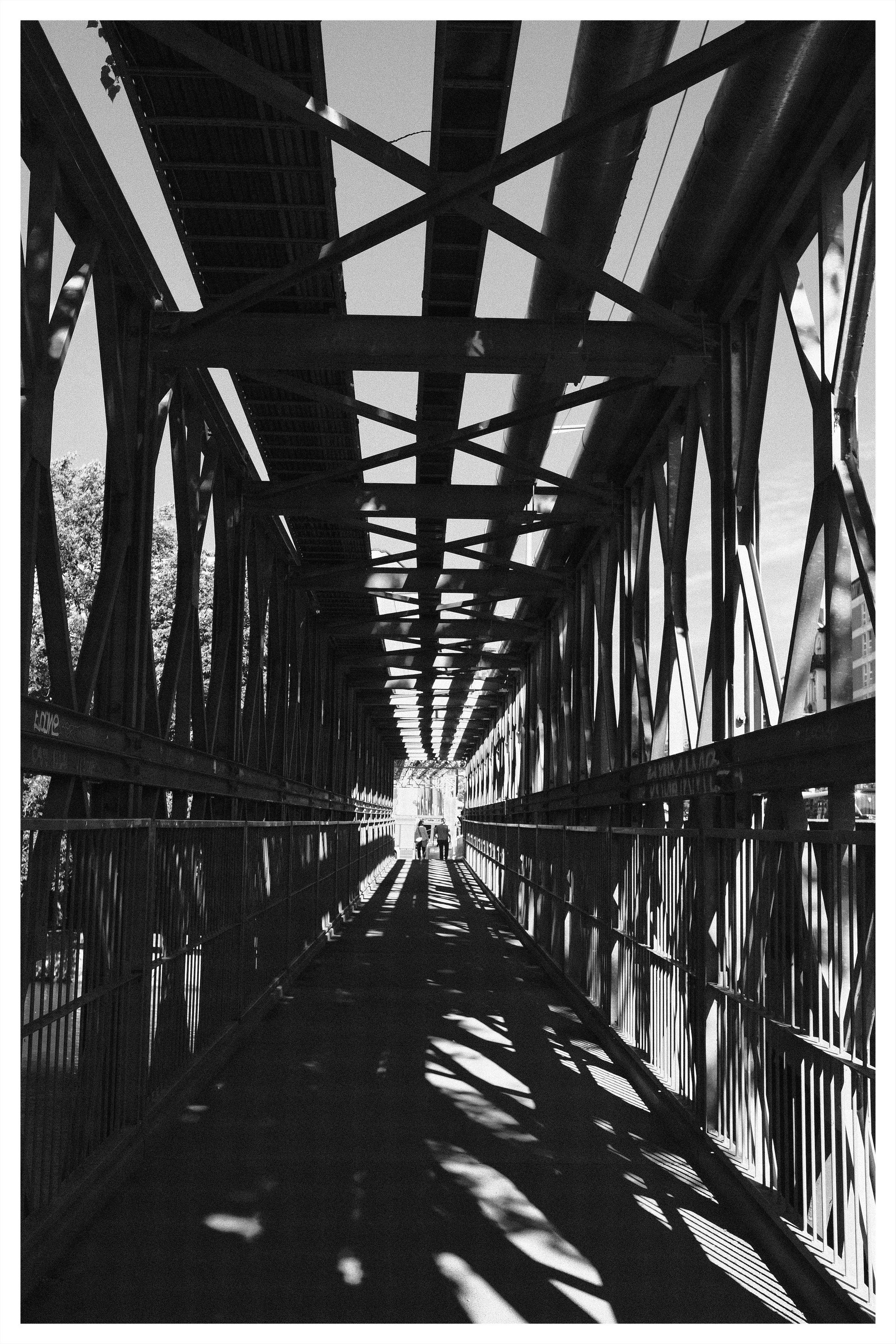 Black-and-white view down a covered steel pedestrian bridge with crisscrossing beams overhead, railings on both sides, and strong shadow patterns on the walkway leading toward a bright opening where a few people are walking.