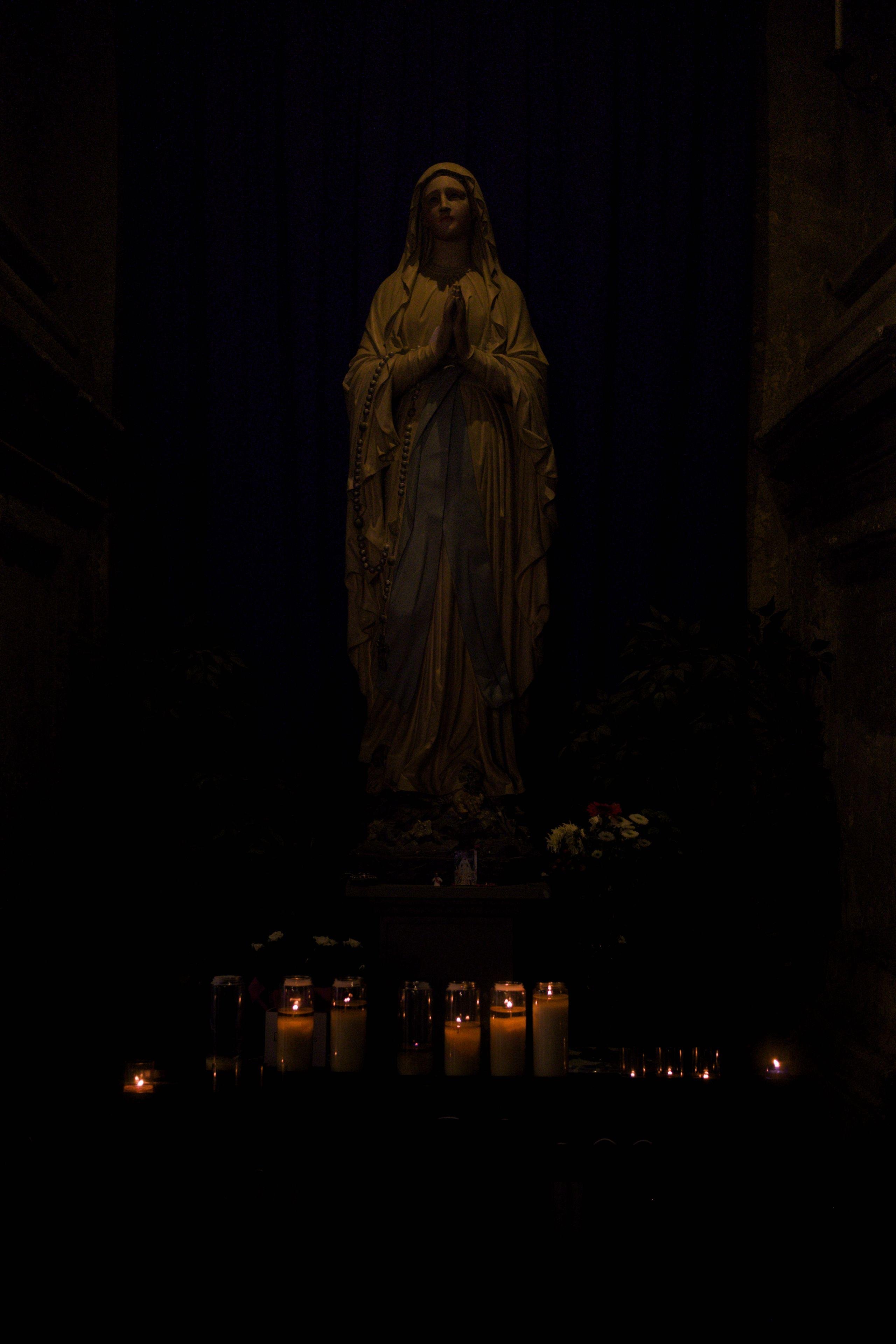 Photo format portrait couleur, montrant une statue blanche de la Sainte Vierge dans une posture classique, mains jointes, priant. Des cierges brûlent à ses pieds, la scène est faiblement éclairée, donnant une ambiance feutrée à l’ensemble