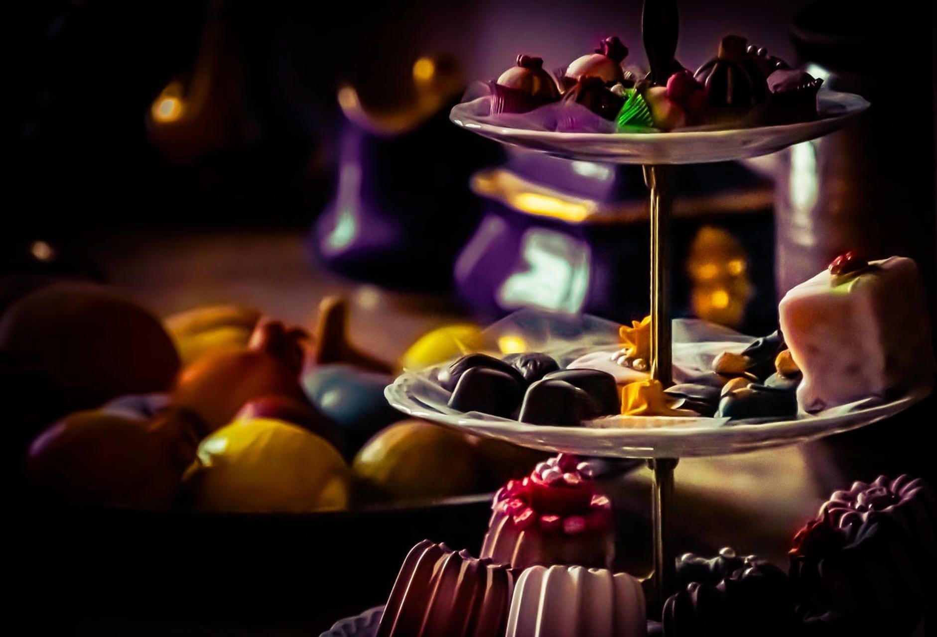 Two-tier glass dessert stand holding assorted chocolates and petit fours, with out-of-focus fruit and purple containers in the dimly lit background.