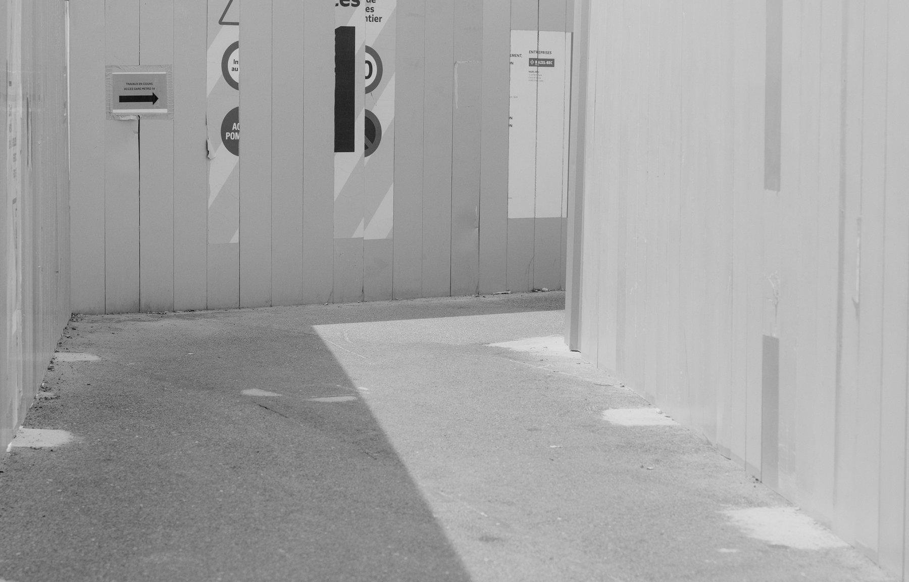 Photo horizontale en noir et blanc. Une allée piétonne entre 2 palissades de travaux grises, le sol aussi est gris faisant perdre nos repères