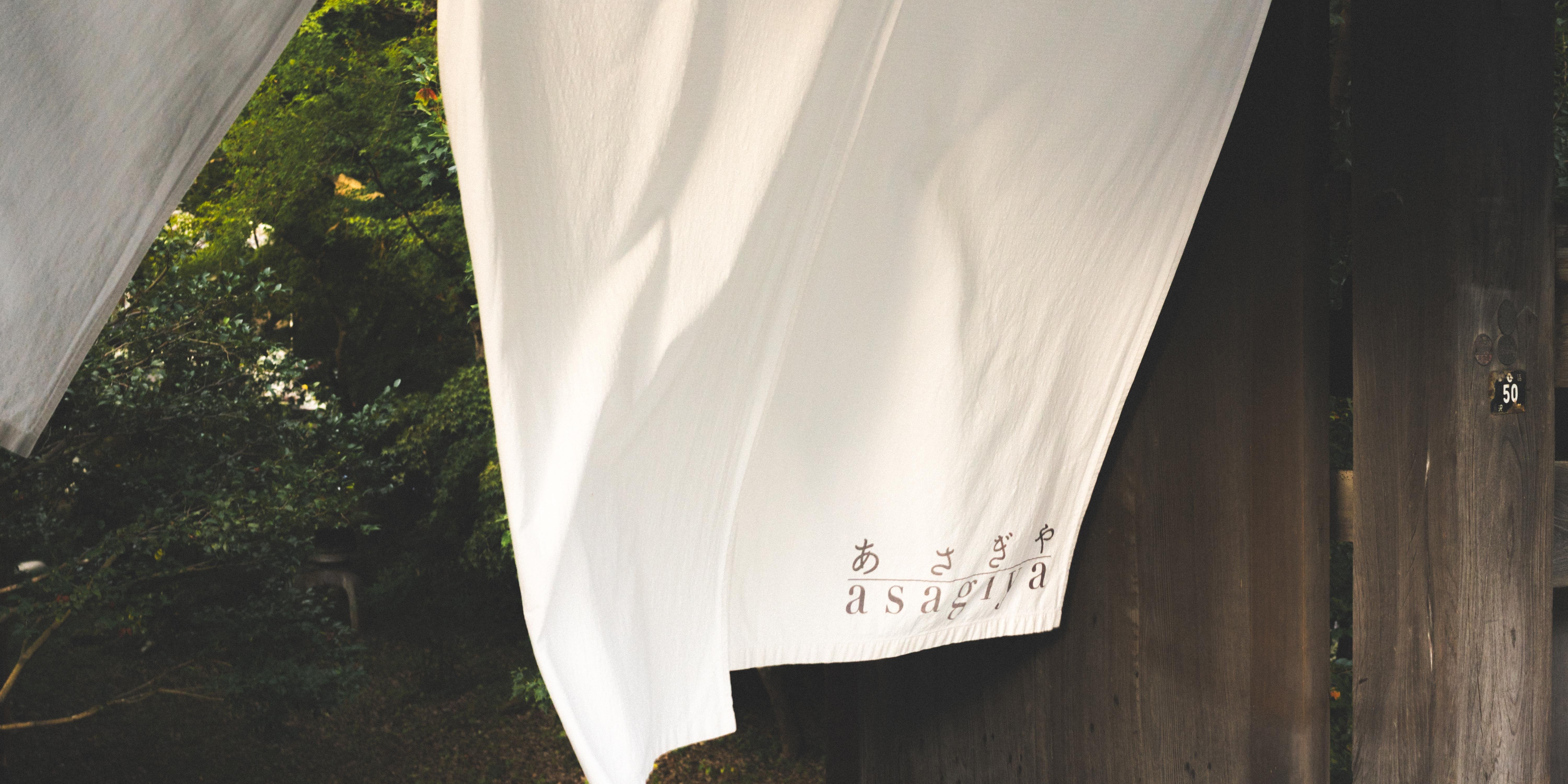 White fabric with Japanese characters and the word "asagura" written near the bottom hangs outdoors. A wooden structure is visible on the right, with a small sign displaying the number 50. Dense, green foliage appears in the background.