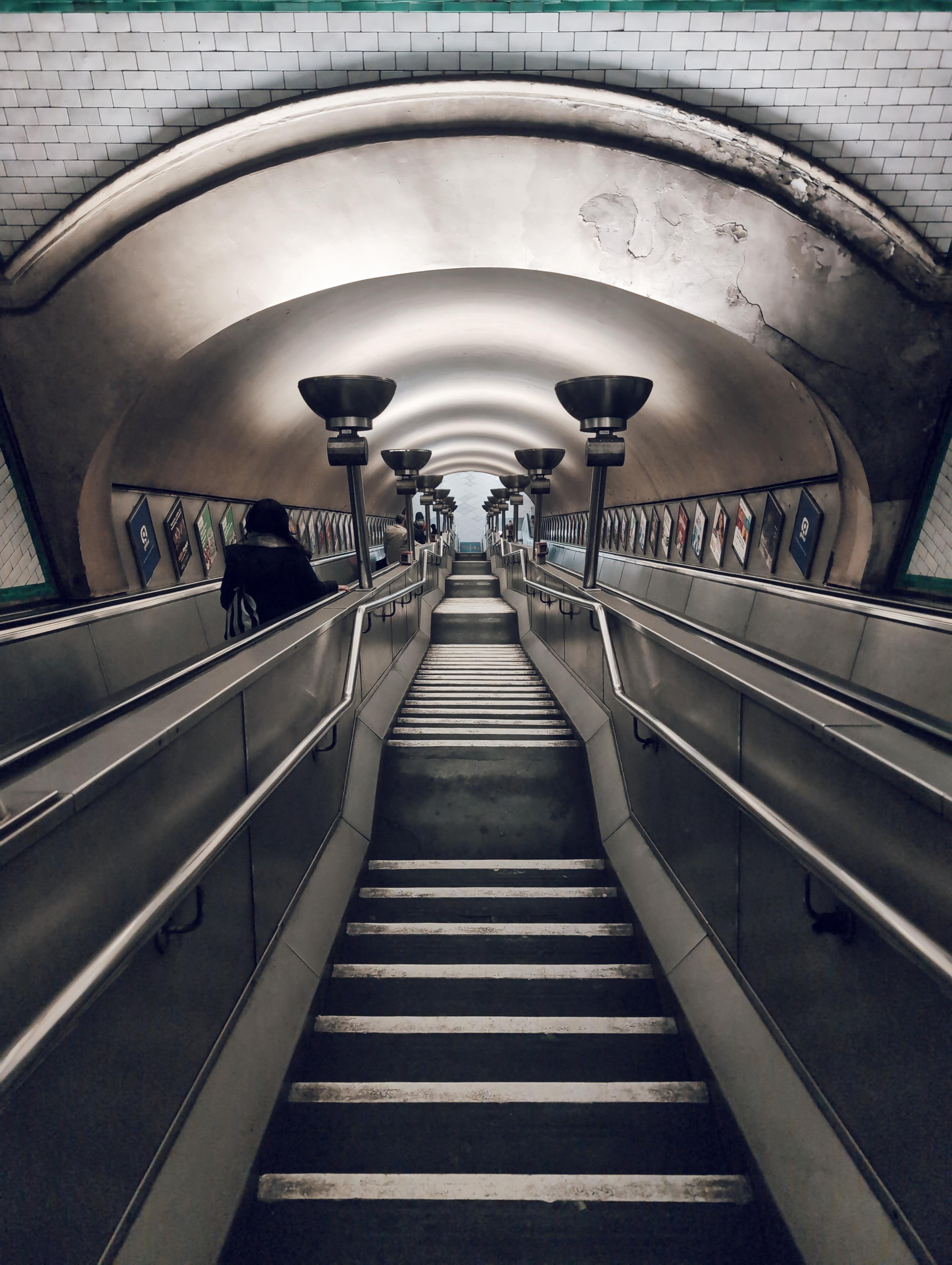 This image shows an underground station escalator with stairs in the middle. The perspective looks upward toward the ceiling, which has an arched, tunnel-like shape. The walls are tiled, and there are lights and advertisements on both sides. There is a person on the left side of the escalator.