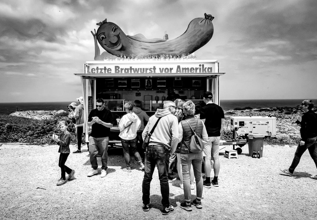 Food truck with a giant sausage decoration and a sign reading "Letzte Bratwurst vor Amerika." Several people stand in front, some purchasing items, others looking at their phones. The background features a cloudy sky and a coastline.