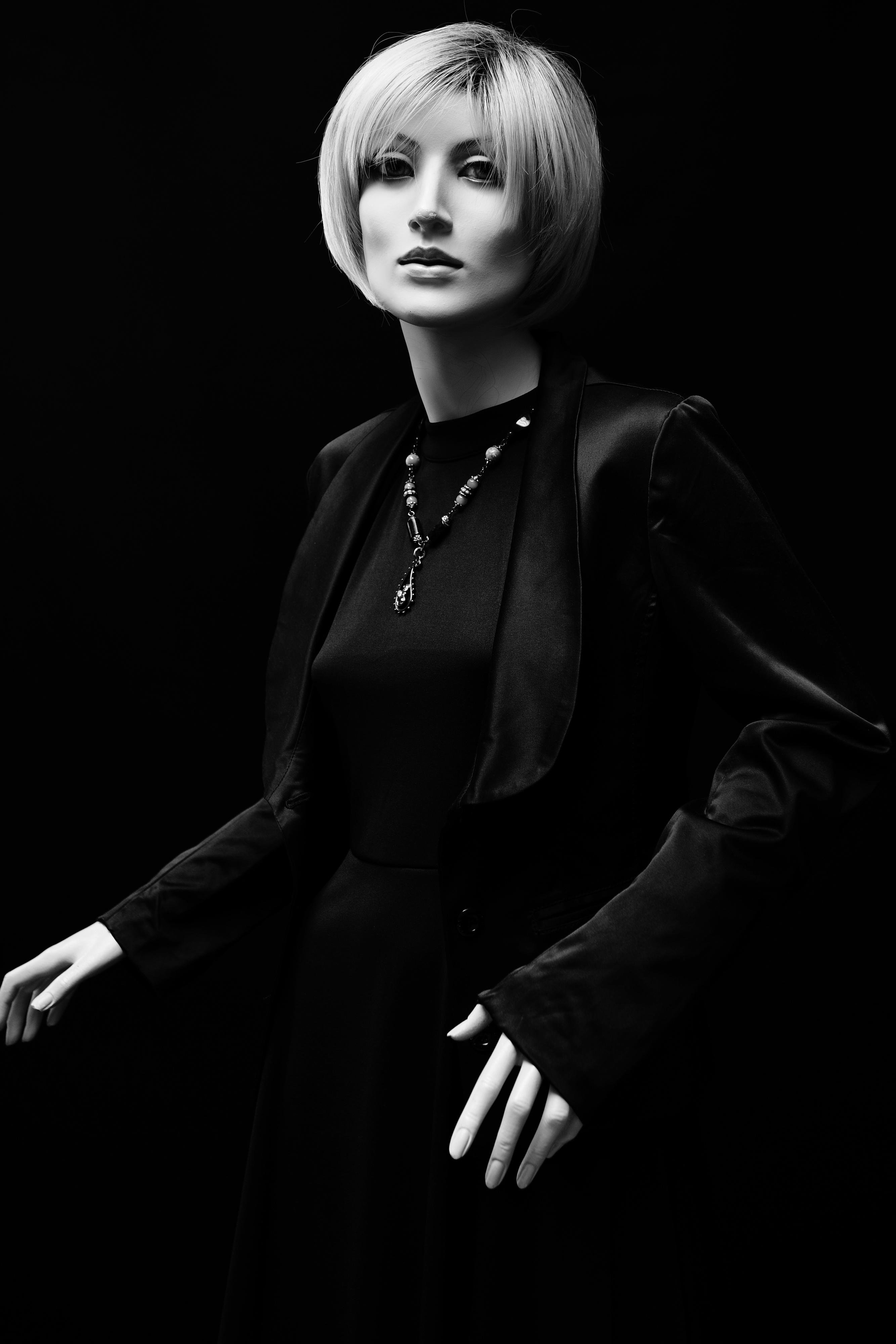 Mannequin with a short, blonde bob haircut dressed in a dark, formal outfit against a black background. Features include a fitted jacket and a necklace with a pendant, adding contrast to the monochrome theme. The pose suggests movement with hands slightly extended.