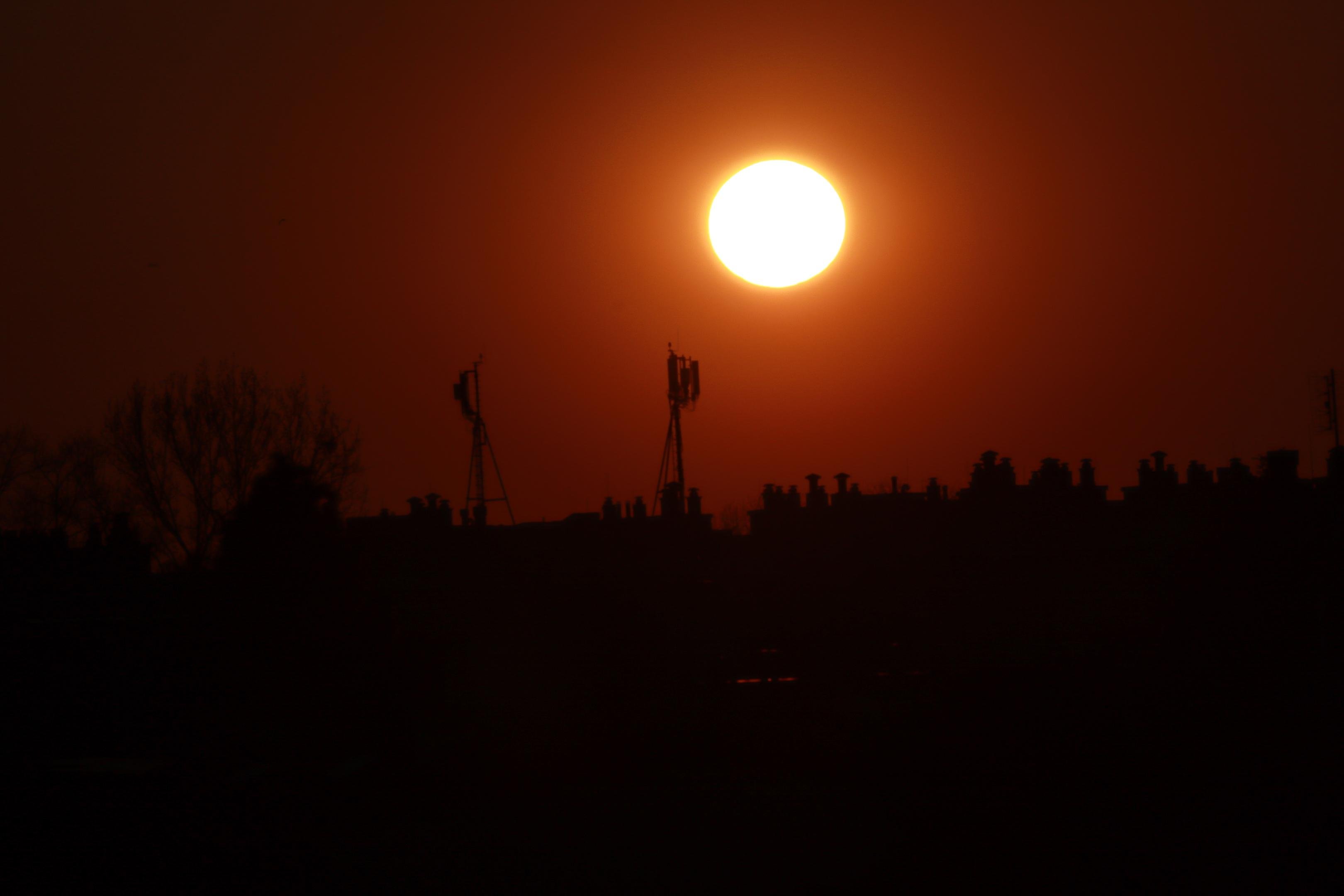 Bright sun low in an orange sky above a dark silhouetted skyline with trees and several tall antenna towers.