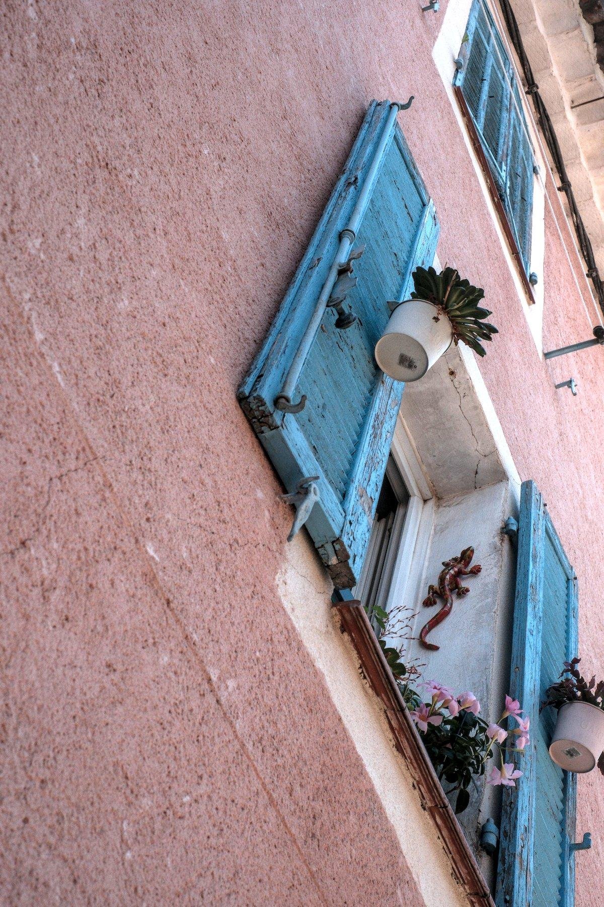 Photo verticale. Contre-plongée vers une fenêtre au volets turquoise ouverts, laissant apparaître une salamandre en céramique accroché au mur blanc, des petits pots de fleur sont accroché sur la face intérieure des volets. Au dessus, la même chose mais volets fermés. Le bâtiment est rose sombre/ocre