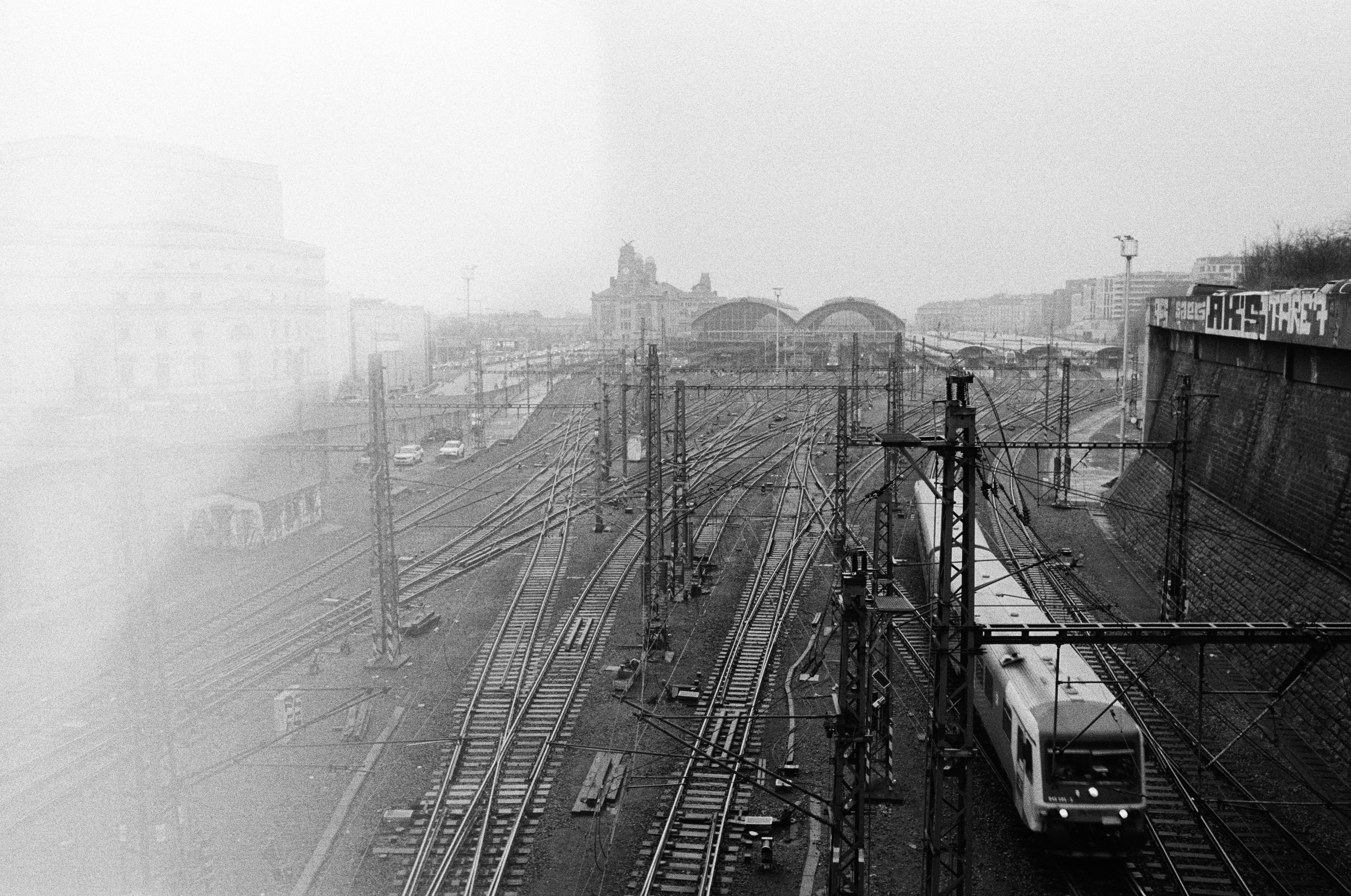 The image shows a black and white scene of a railway station with multiple train tracks converging towards the station in the background. There is a train on one of the tracks to the right. Overhead electrical lines and supporting poles are visible. Buildings can be seen in the distance, and there is graffiti on a wall to the right. There is a significant light damage on the left.
