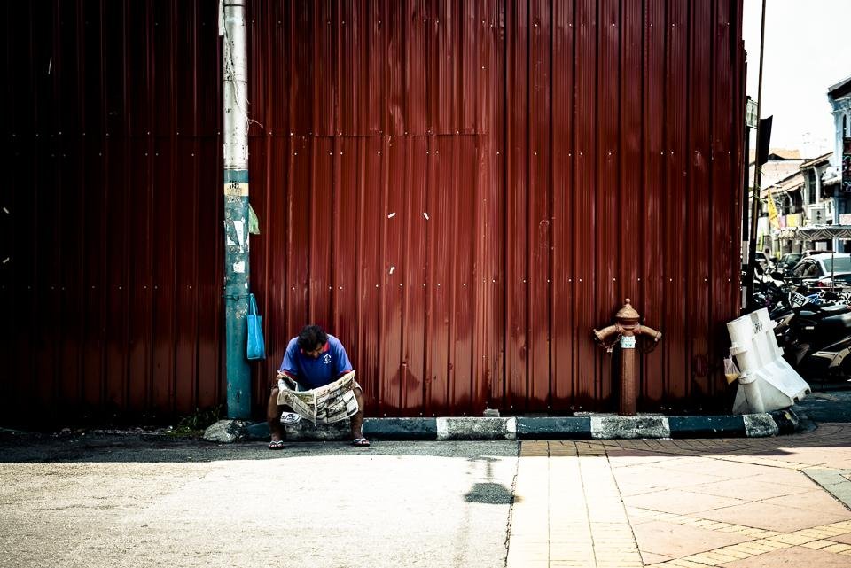 Person sitting on a curb reading a newspaper beside a large red corrugated metal wall, with a utility pole to the left, a fire hydrant near the center-right, and parked motorcycles lining the street in the background.