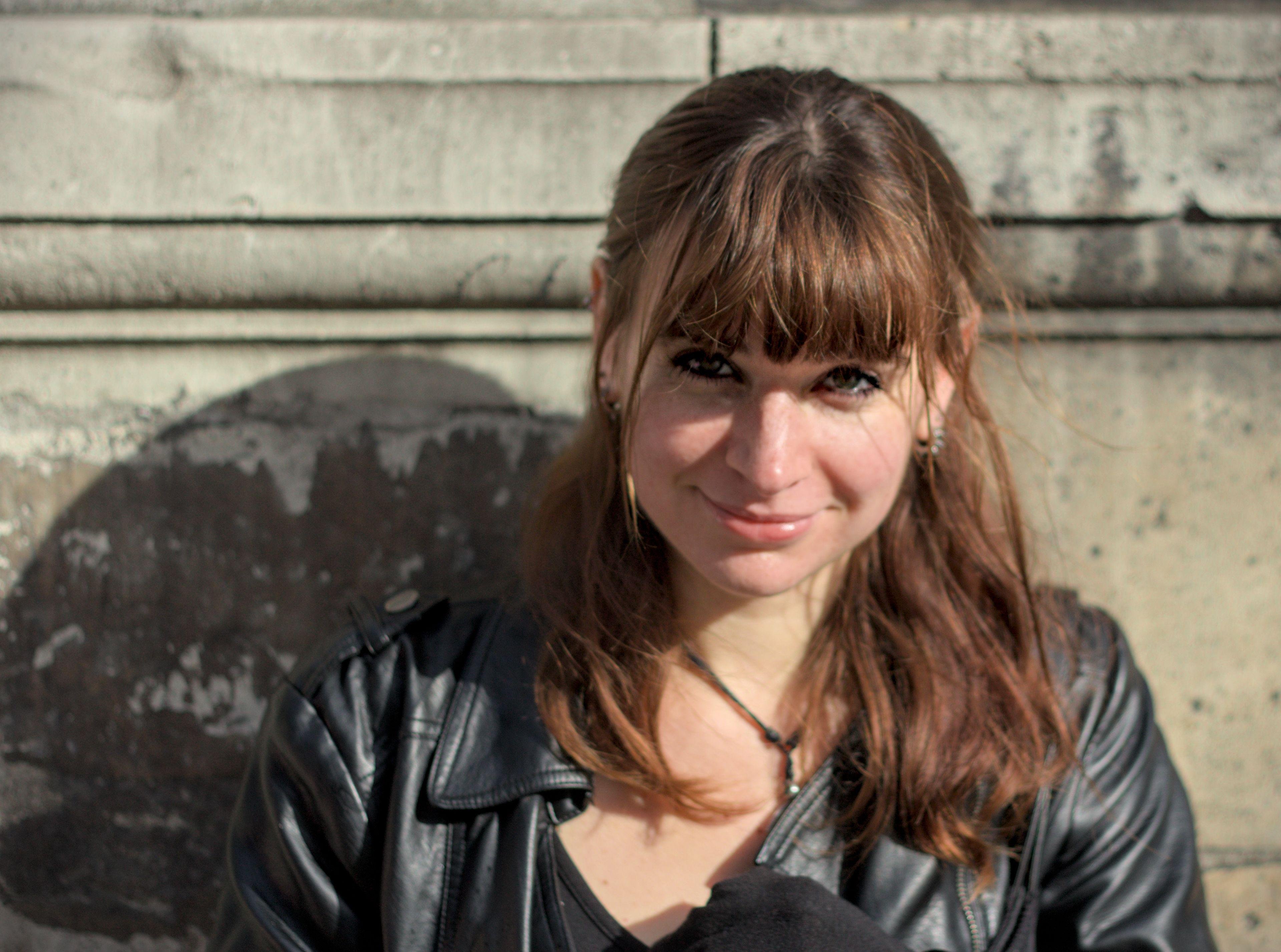 Photo format paysage montrant une jeune femme de face, appuyée contre un mur dehors, éclairée pas le soleil. Elle porte un blouson en cuir et regarde la caméra intensément en souriant