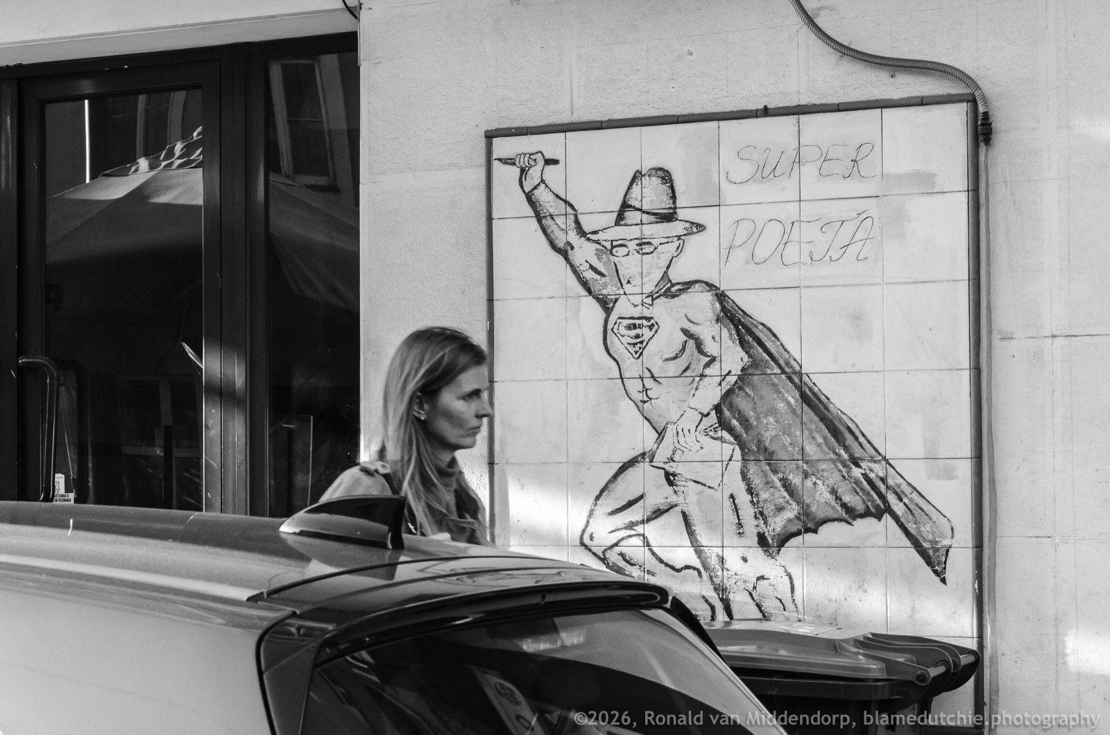 Black-and-white street scene with a woman walking beside a parked car in the foreground; behind her, a tiled wall features tiles with Fernando Pessoa as superhero with the words “SUPER POETA,” next to a window and doorway.