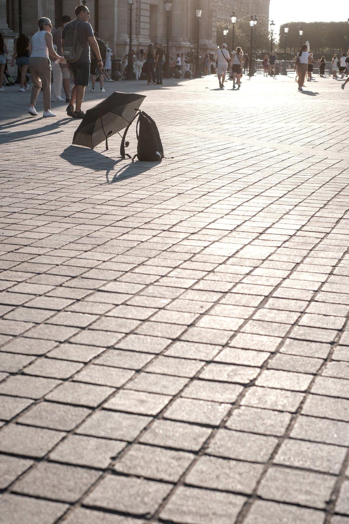 Photo verticale. Vue assez bas sur les pavés de la cours du Louvre, vides jusqu’en haut de l’image où un sac à dos et un parapluie ouvert sont posés au sol. Des gens passent à côté sans y prêter attention, le soleil rasant arrive d’en haut à droite