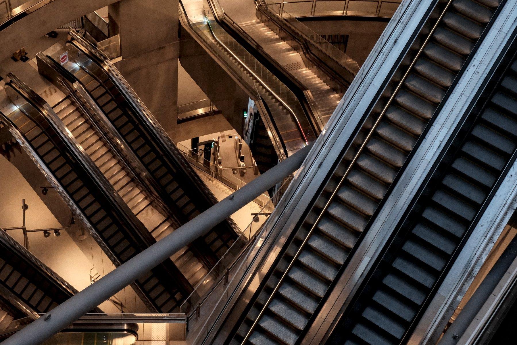 Photo horizontale. Plongée sur des escalators qui descente dans les profondeurs d’une grande station. Les plus proches de la caméra suivent un axe SW-NE, et réfléchissent la lumière du jour, un peu bleutée, les plus profonds suivent un axe NW-SE et réfléchissent l’éclairage artificiel orangé