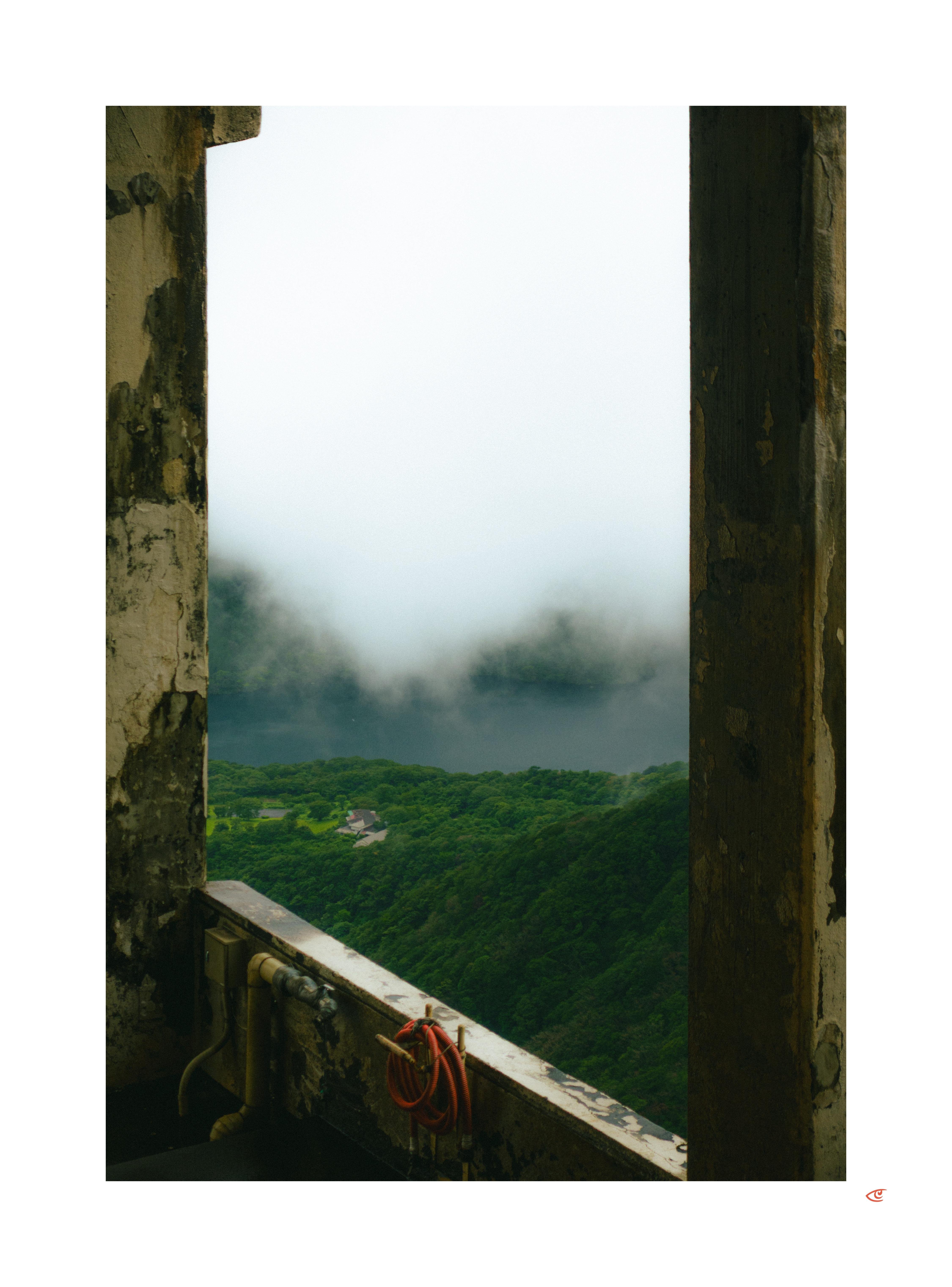 View through a tall rectangular window opening in a weathered concrete wall, showing a green forested hillside with a few small buildings below and low clouds or fog in the distance; a red coiled hose hangs on the inner ledge near exposed pipes.