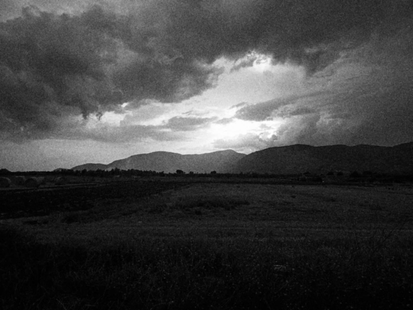 Paesaggio di campagna al tramonto, con balle di fieno, montagne in lontananza e cielo nuvoloso.