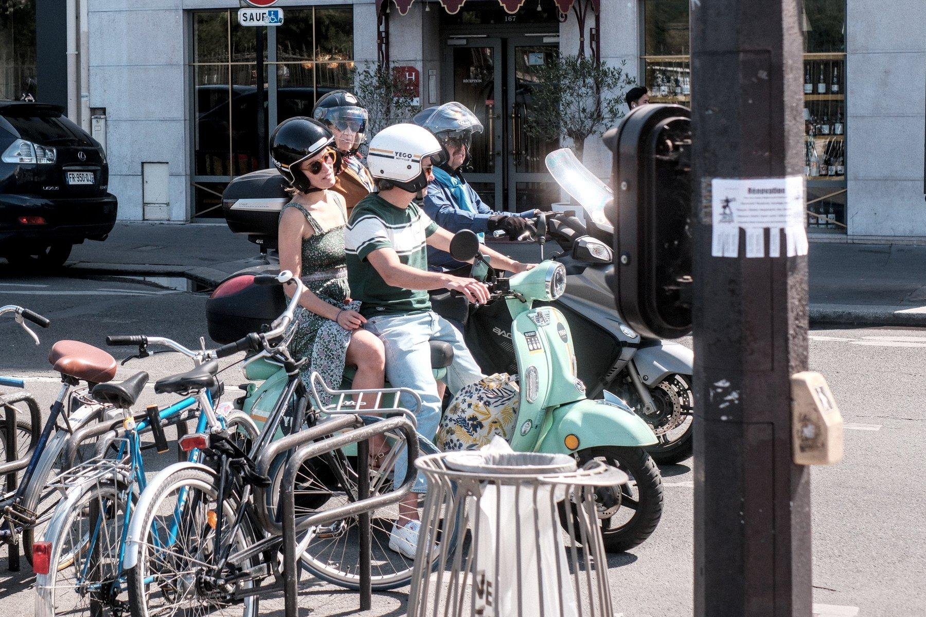 Photo horizontale. Arrêté à un feu rouge, tournés vers la droite, un vespa vert pâle conduit par un homme en jean, t-shirt vert avec une grande bande horizontale blanche au niveau de la poitrine, casque blanc et derrière lui, une femme en passagère, robe à bretelle verte au motifs floraux, lunettes de soleil, casque noir. à leur gauche (derrière de notre point de vue, une moto moderne noire, conduite par un homme à moustache et lunette de soleil, casque noir avec une passagère  derrière aussi, casque noir, lunette de soleil et veston orange