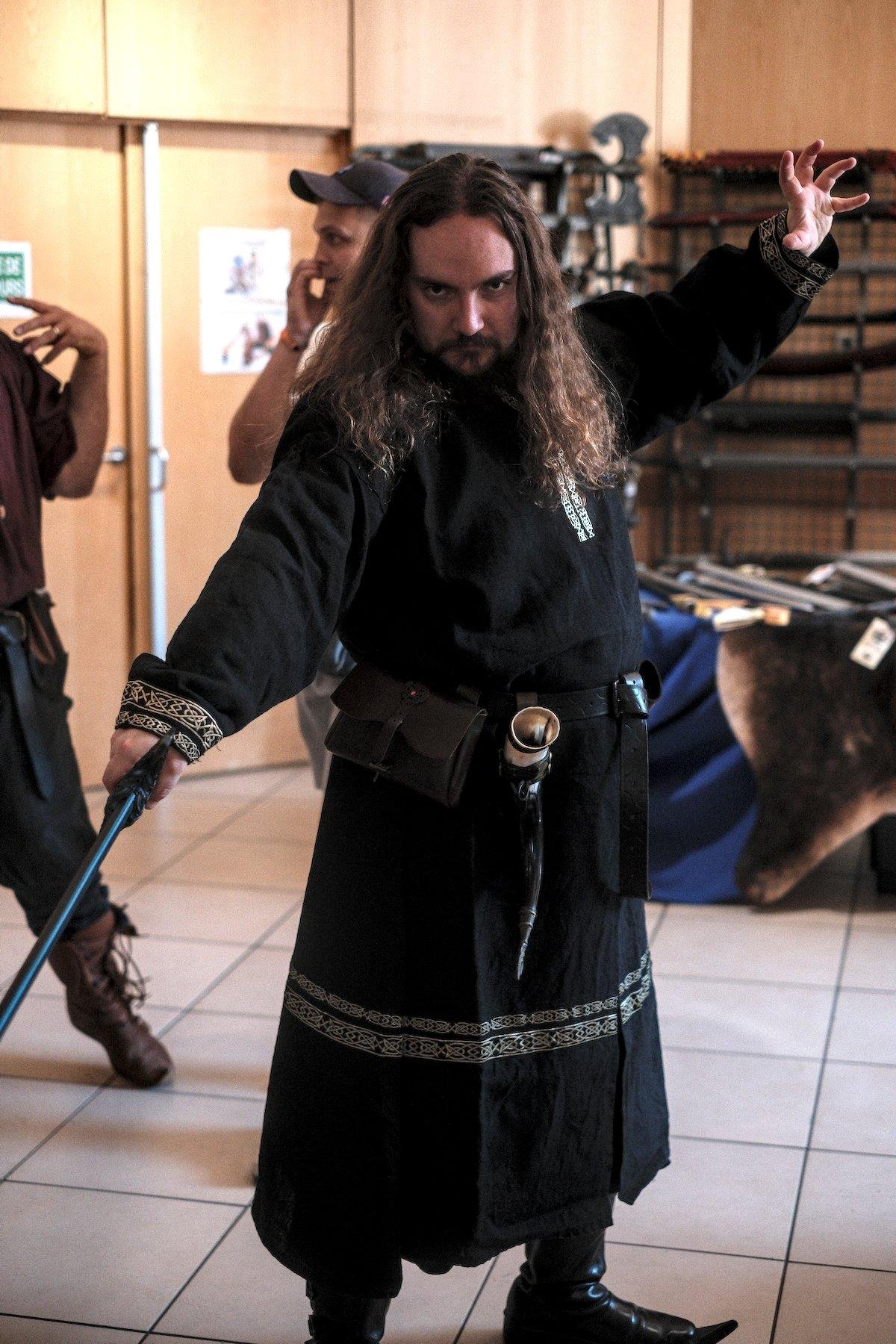 Photo verticale. Portrait de plein pied d’un homme en costume heroic fantasy, une longe toge noire avec des décoration argenté brodées en motif croisés évoquant les Celtes, une ceinture en cuir avec une sacoche en cuir, une corne de brume attachée. Il prend une pose menaçante avec son épée main droite, et la main gauche crispée en l’air derrière lui comme si il concentrait de la magie