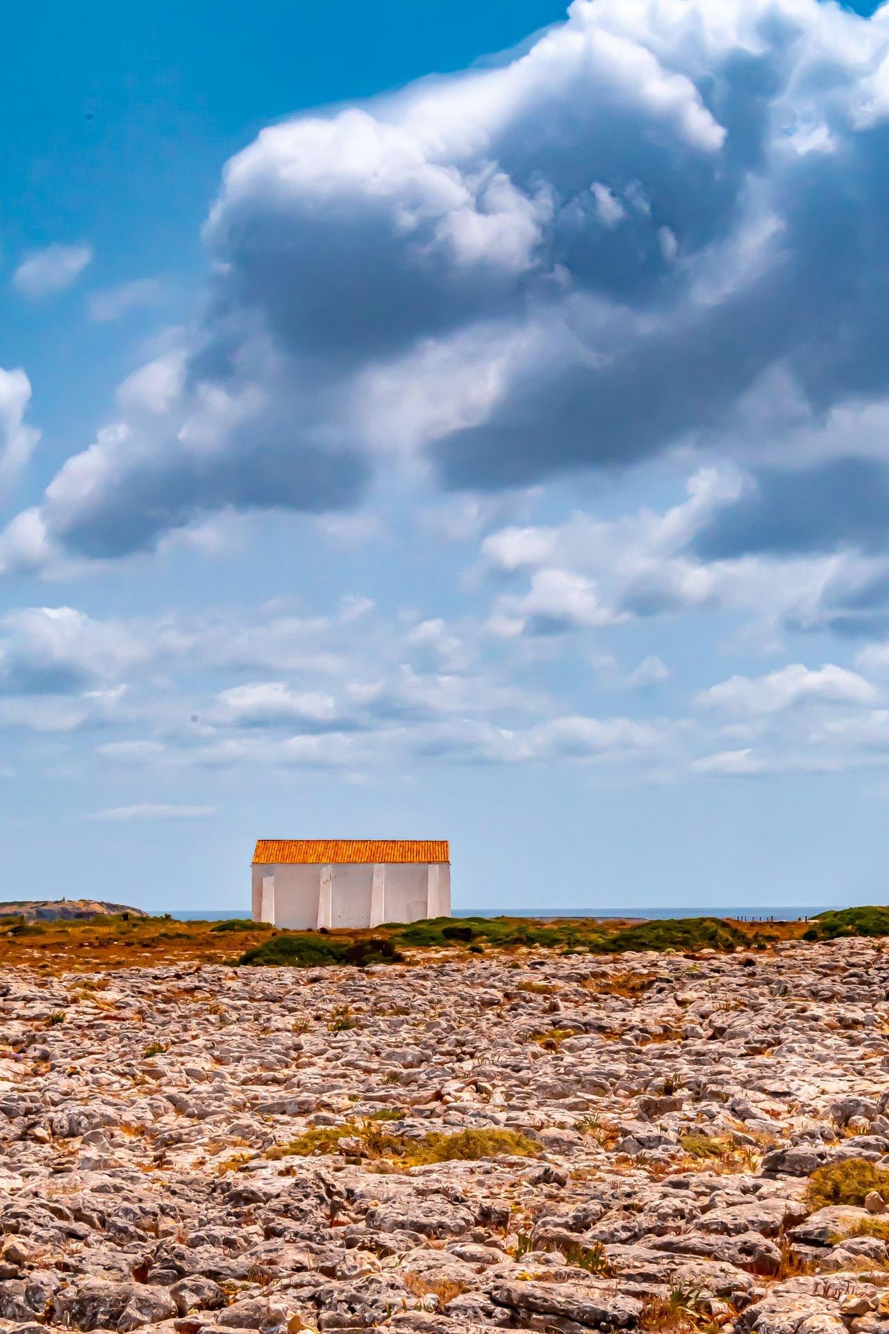 White building with a red-orange roof sits alone on a rocky landscape under a blue sky filled with large, fluffy clouds. Vegetation is sparse, with some green bushes scattered around. The horizon in the background suggests a coastal area with the sea visible.
