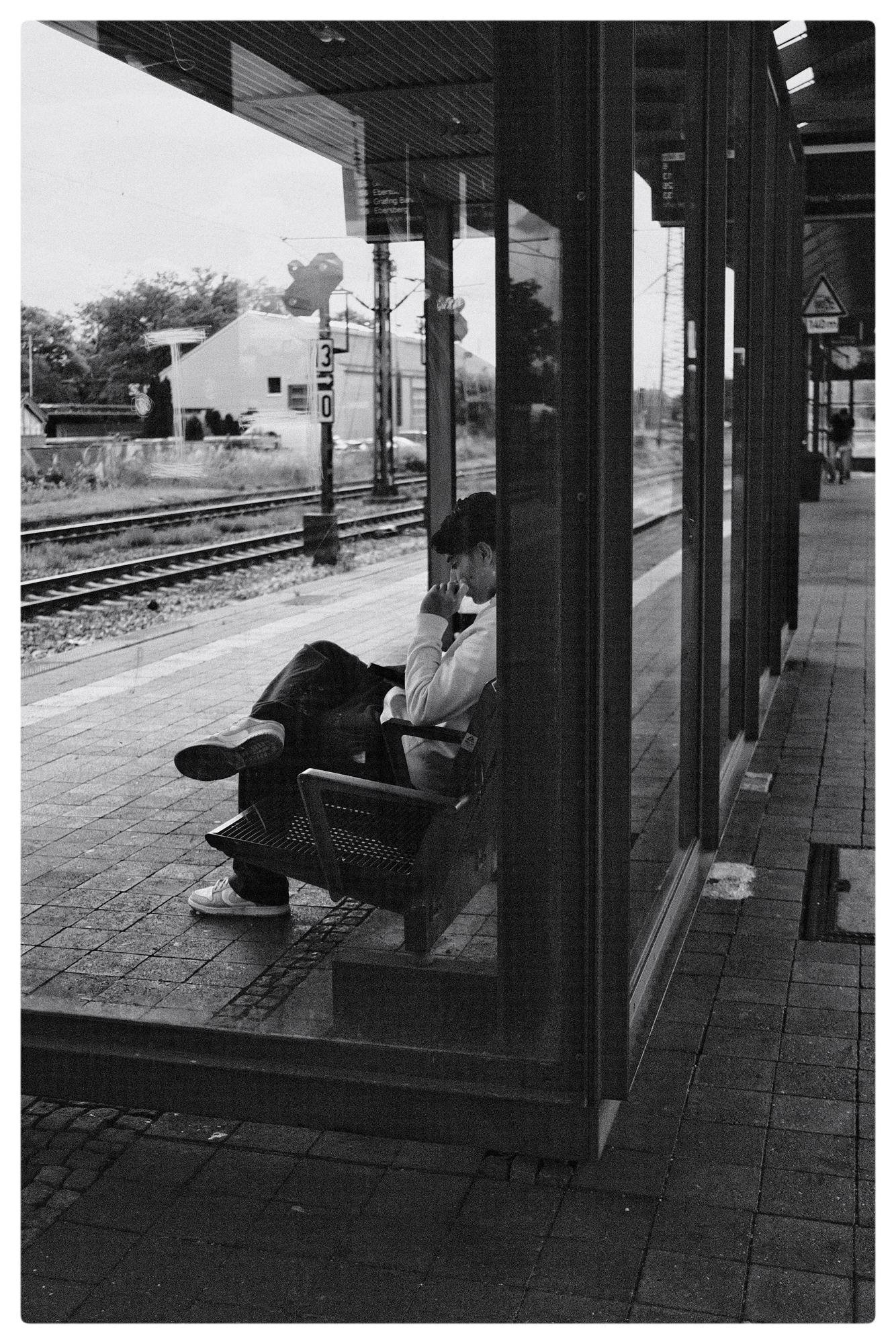 Person sitting on a bench in a covered train station platform, wearing a jacket and cap, with legs crossed. The platform is empty except for the seated figure, with visible train tracks and a small building in the background. The scene appears in black and white.