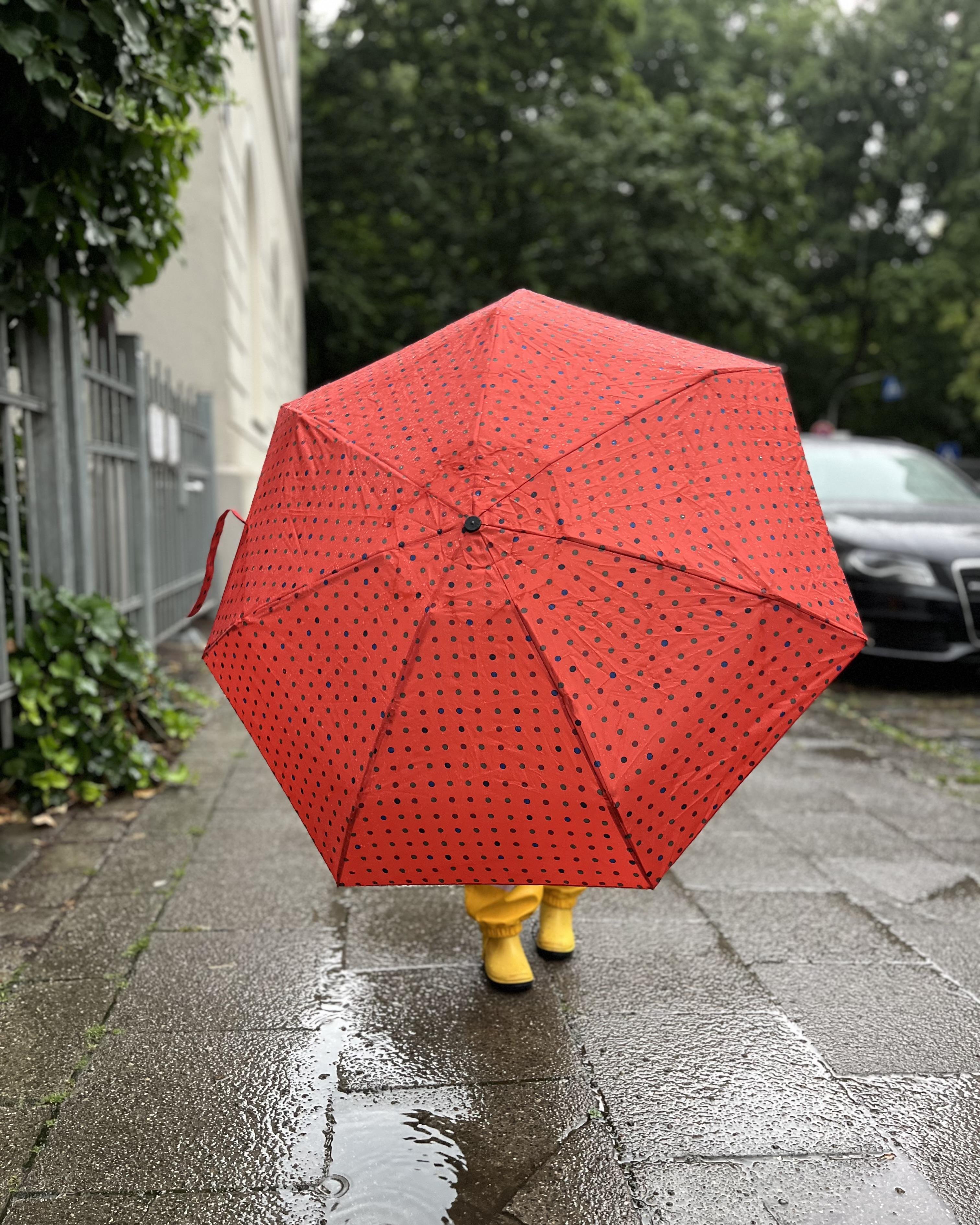 Rather close-up wide angle shot of a child on a rainy street holding a red umbrella with small black dots. The toddler points the opened umbrella right towards the camera, so that only the yellow rain boots of the child can be seen below the umbrella. The bright colors of the yellow boots and the red umbrella contrast the grey city scene in the background.