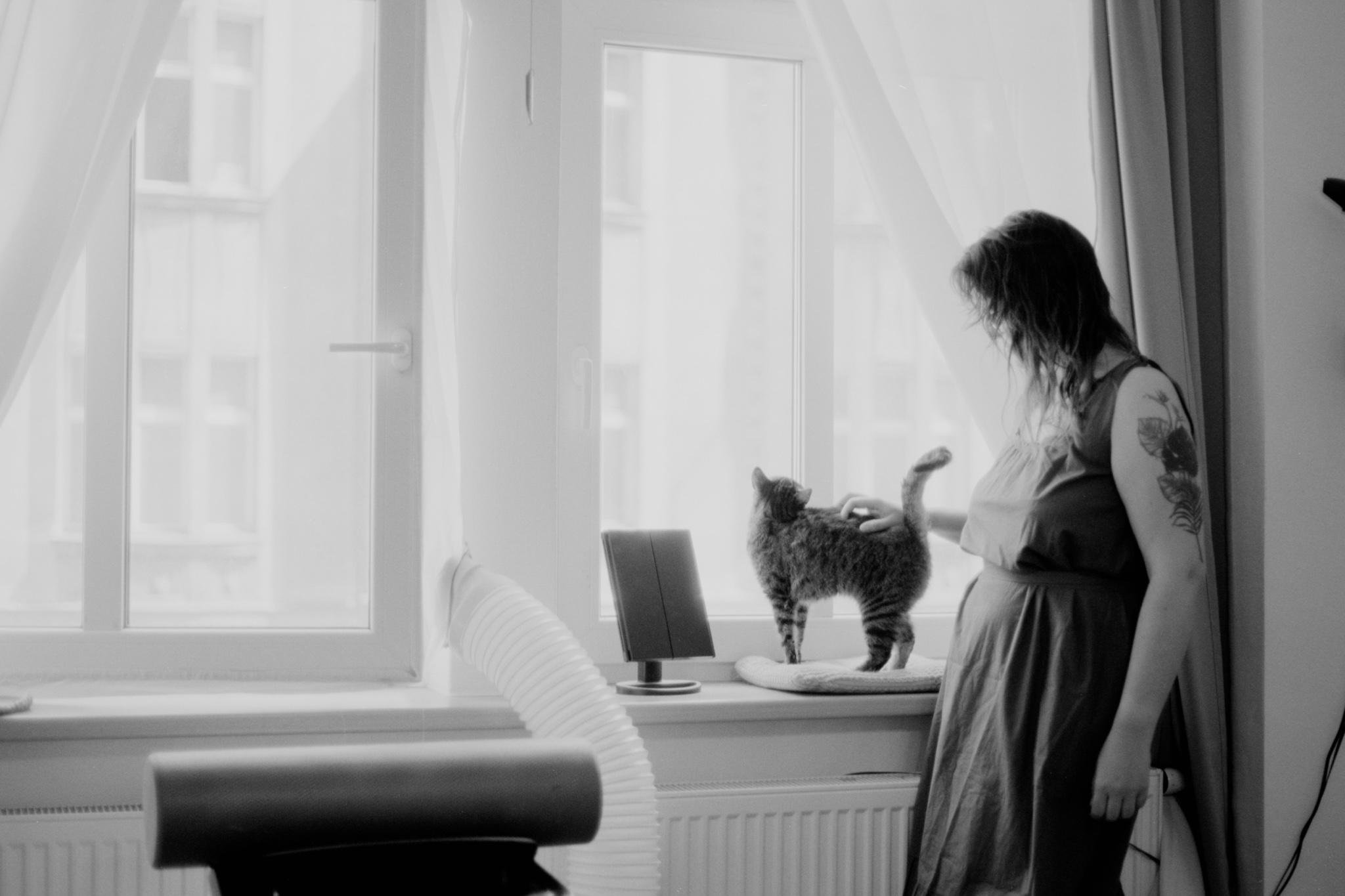 Black-and-white photo of a woman with a tattooed arm standing by a window, petting a cat on the windowsill beside a small table mirror, with sheer curtains and a vent hose in the foreground.