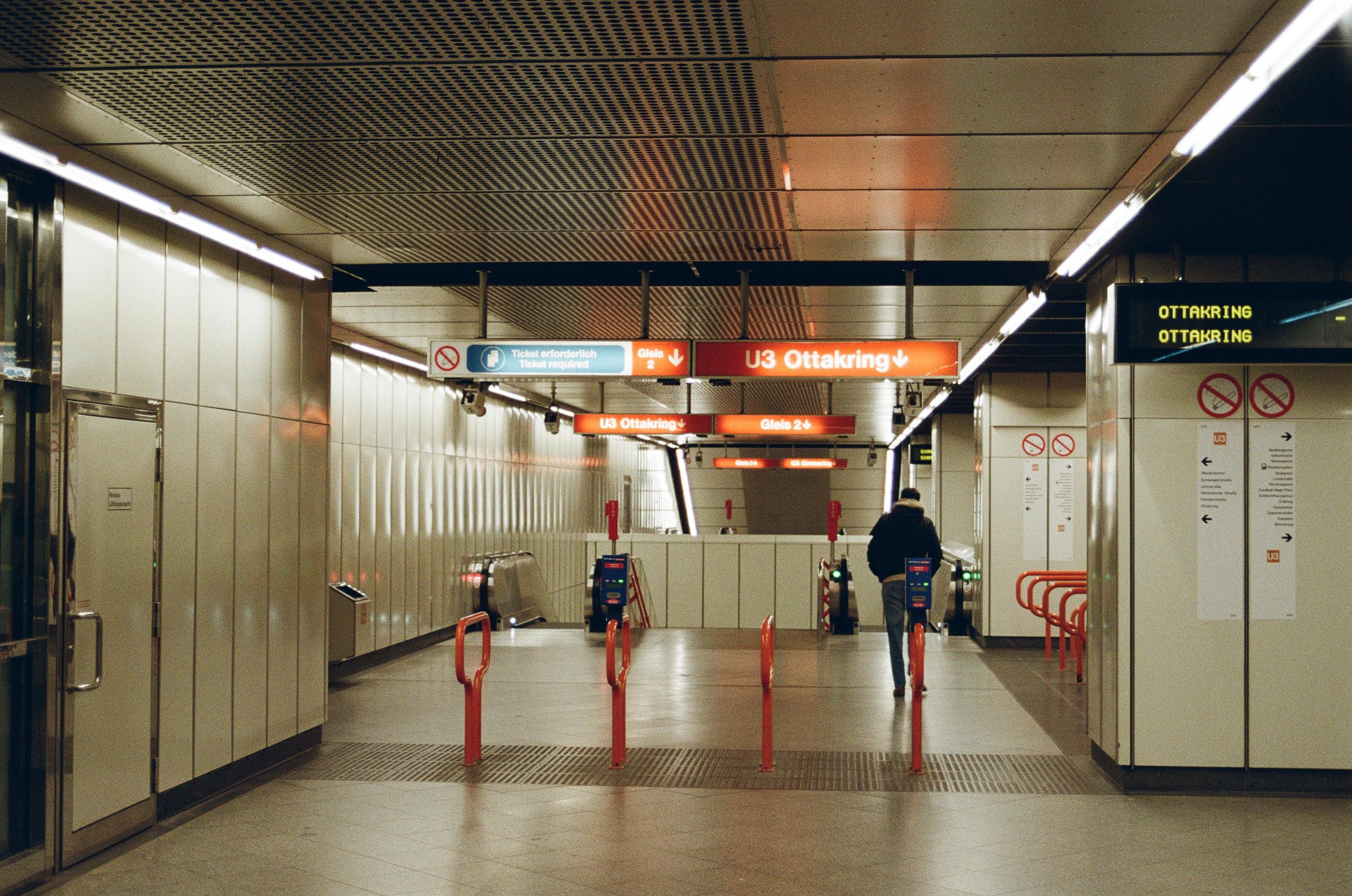 A photo of a metro station. It features signage indicating the U3 line towards Ottakring, and there are red barriers along a walkway leading to escalators. The station has a modern, clean appearance with overhead lighting and informational signage. There is a person walking towards the escalators.