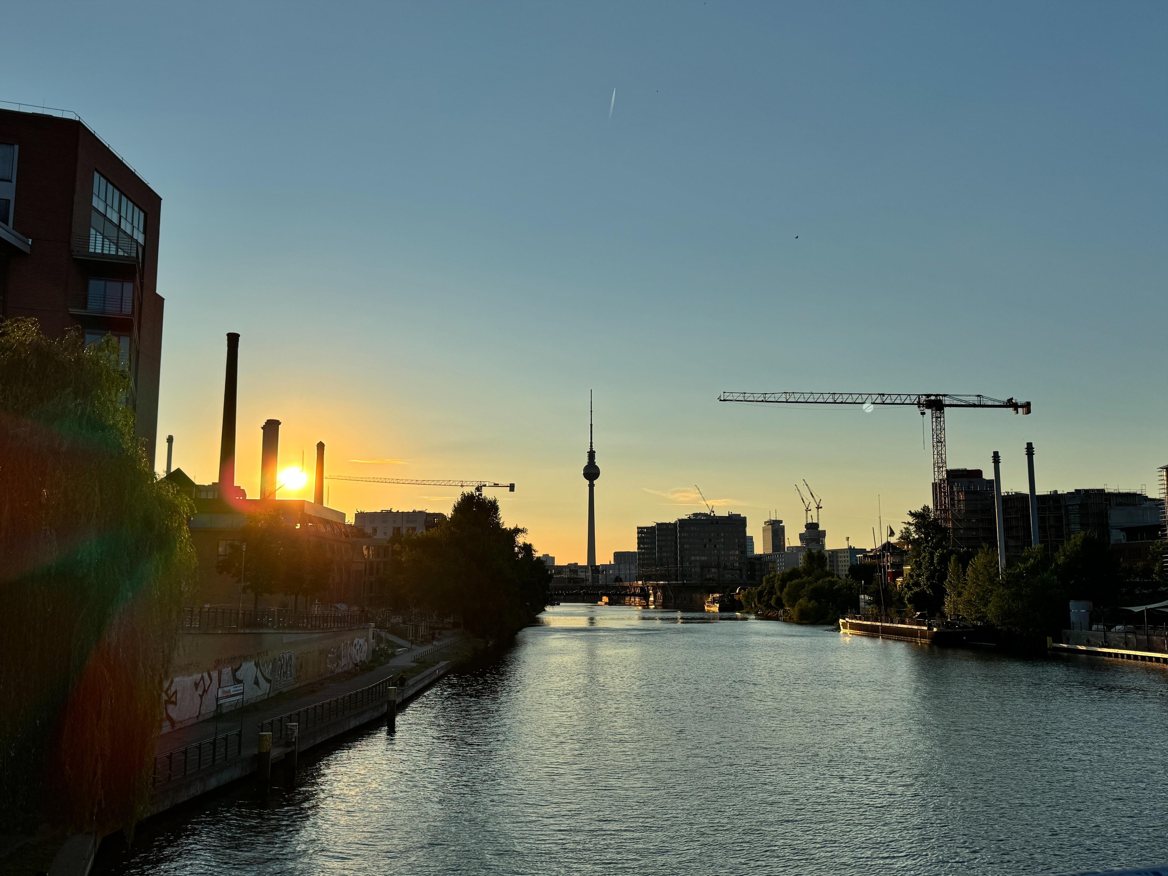 A river flows between buildings with construction cranes and chimneys under a sunset sky. The Berlin TV Tower is visible in the distance.
