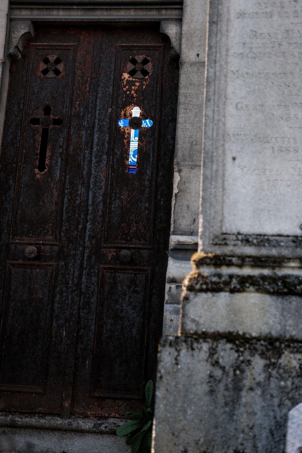 Photo verticale. Une porte de mausolée totalement rouillée, avec des ouvertures en forme de croix, dont l’une est alignée avec le vitrail au fond du mausolée, formant une croix de lumière colorée