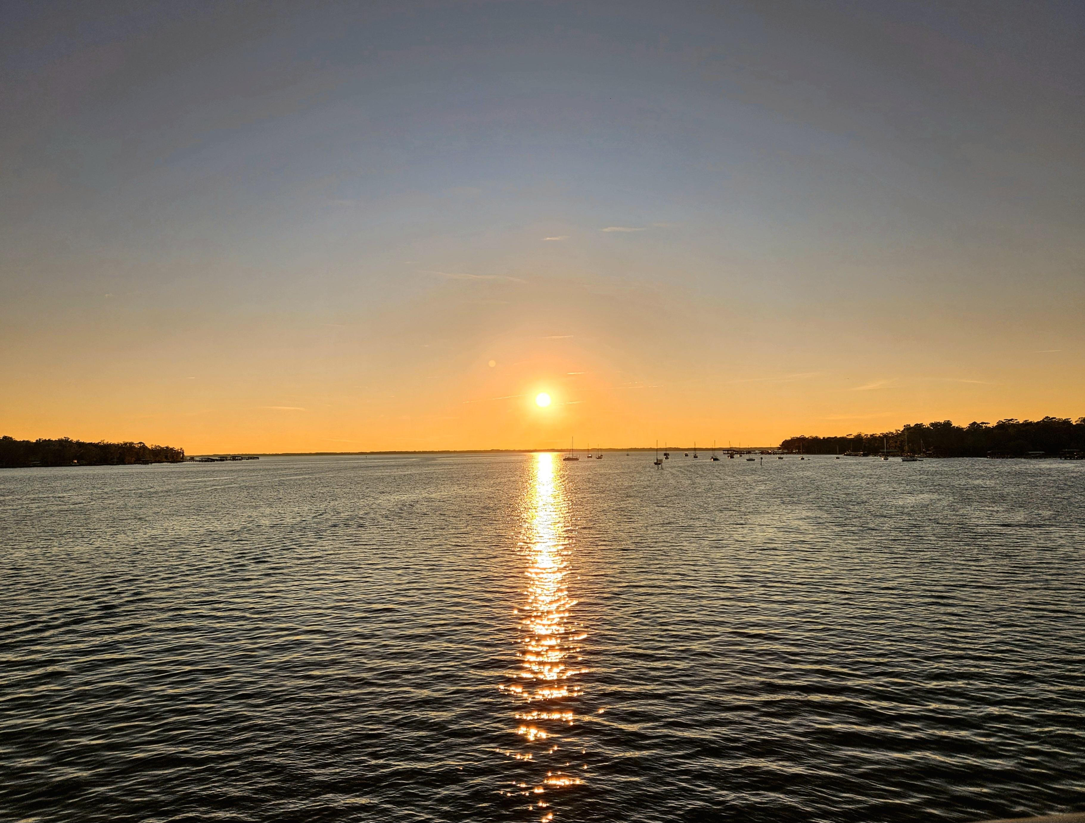 Sun setting over a large body of water, casting a bright, reflective trail across the surface. The sky transitions from blue to warm orange hues near the horizon. Silhouetted trees line both sides of the horizon, with scattered small boats visible in the distance.