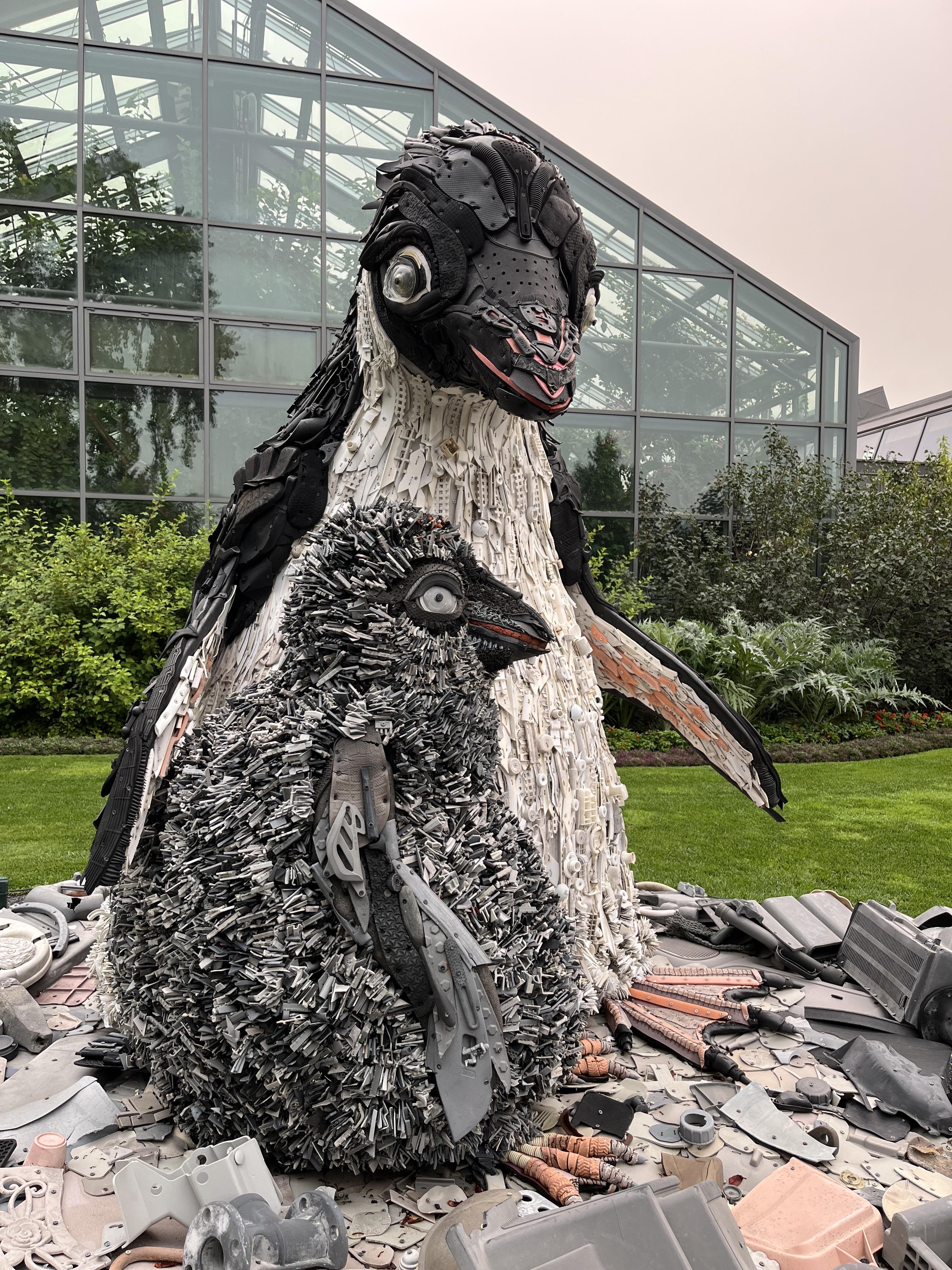 Two penguin sculptures made from recycled materials, with one adult and one chick. The adult penguin has a body covered in white and black plastic components, while the chick has a textured surface resembling metal shavings. Both stand on a circular base with scattered industrial materials. The background features a greenhouse with large glass panels and surrounding greenery.