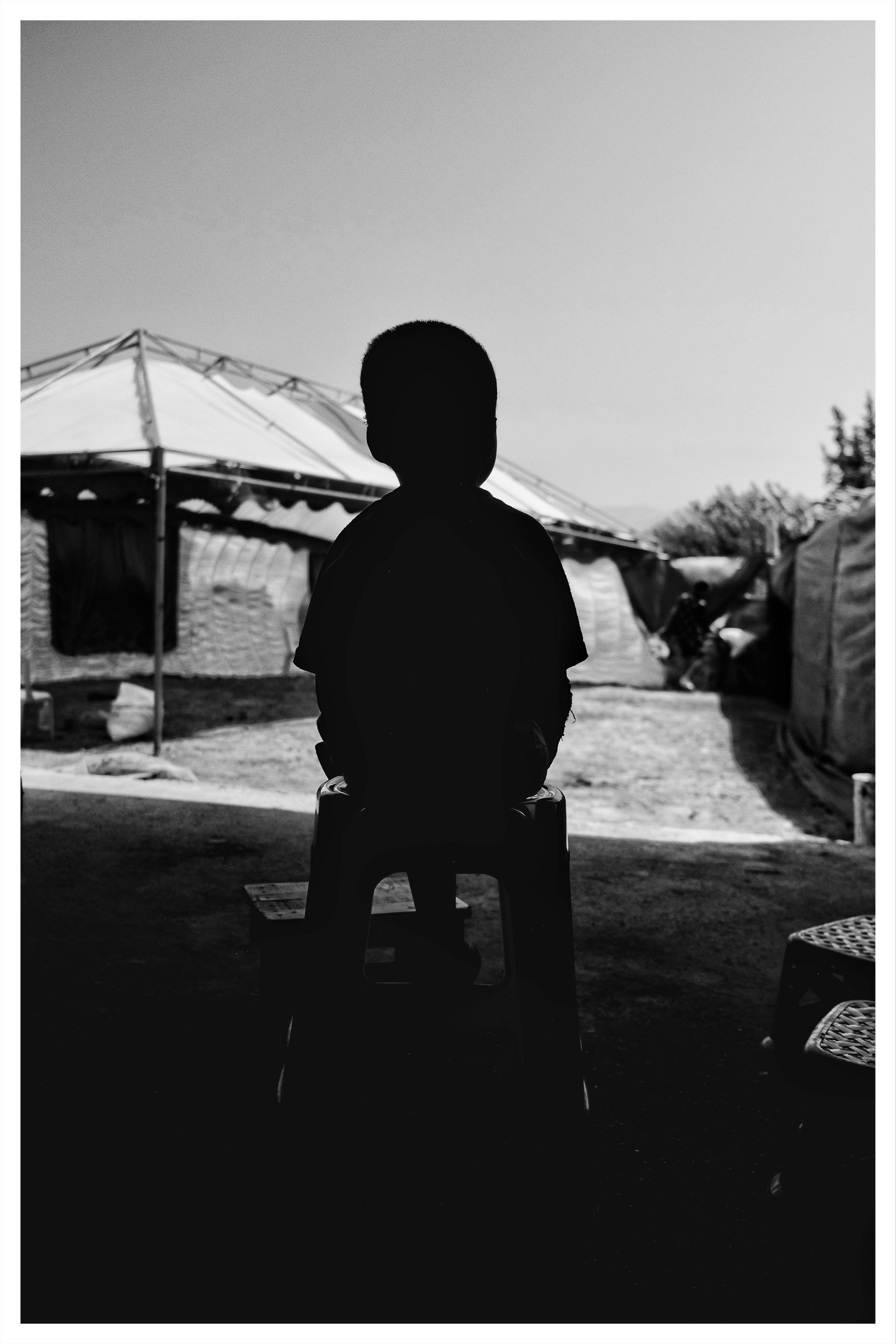 Silhouette of a child sitting on a small plastic stool, centered in the foreground, with a canopy tent and outdoor campsite structures in the background under a bright sky.