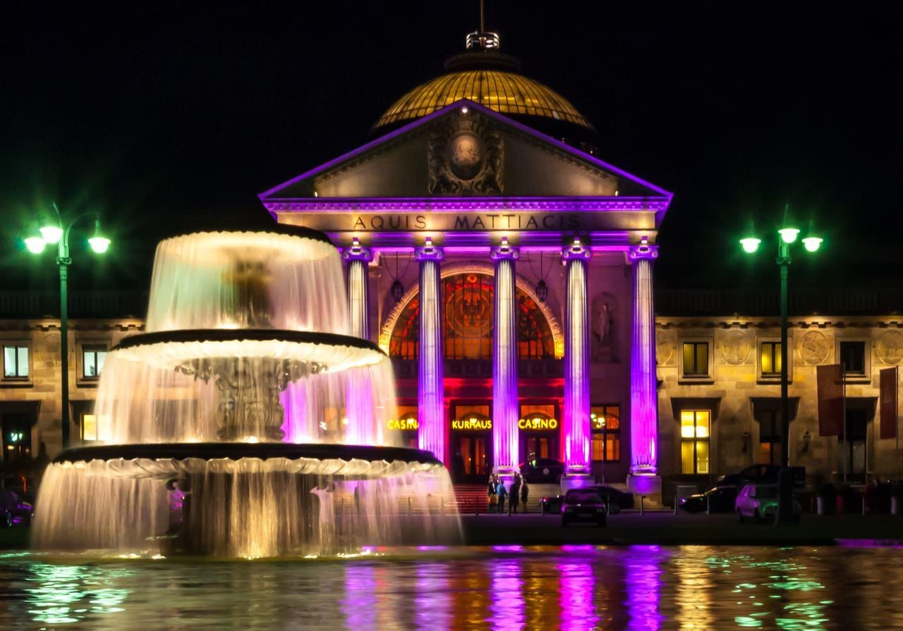 The image shows a well-lit building at night, featuring a dome and columns, with the words "AQUIS MATTACIS" on the facade. In front of the building is an ornate, multi-tiered fountain with water flowing down. The lighting includes vibrant colors like purple, giving it a festive atmosphere.