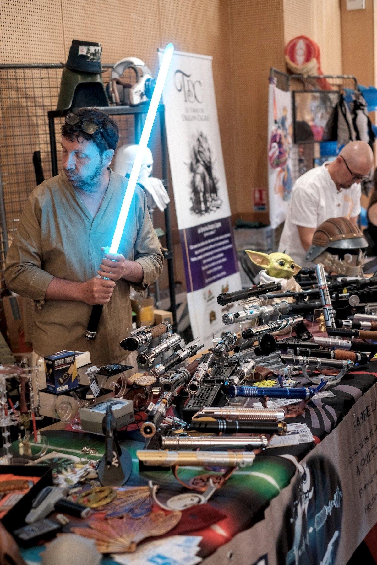Photo verticale. Un stand dans une convention, avec sur la table, plein de sable laser différents. Derrière la table, le tenancier avec un sabre laser allumé, bleu, et écoutant quelqu’un hors cadre sur la gauche