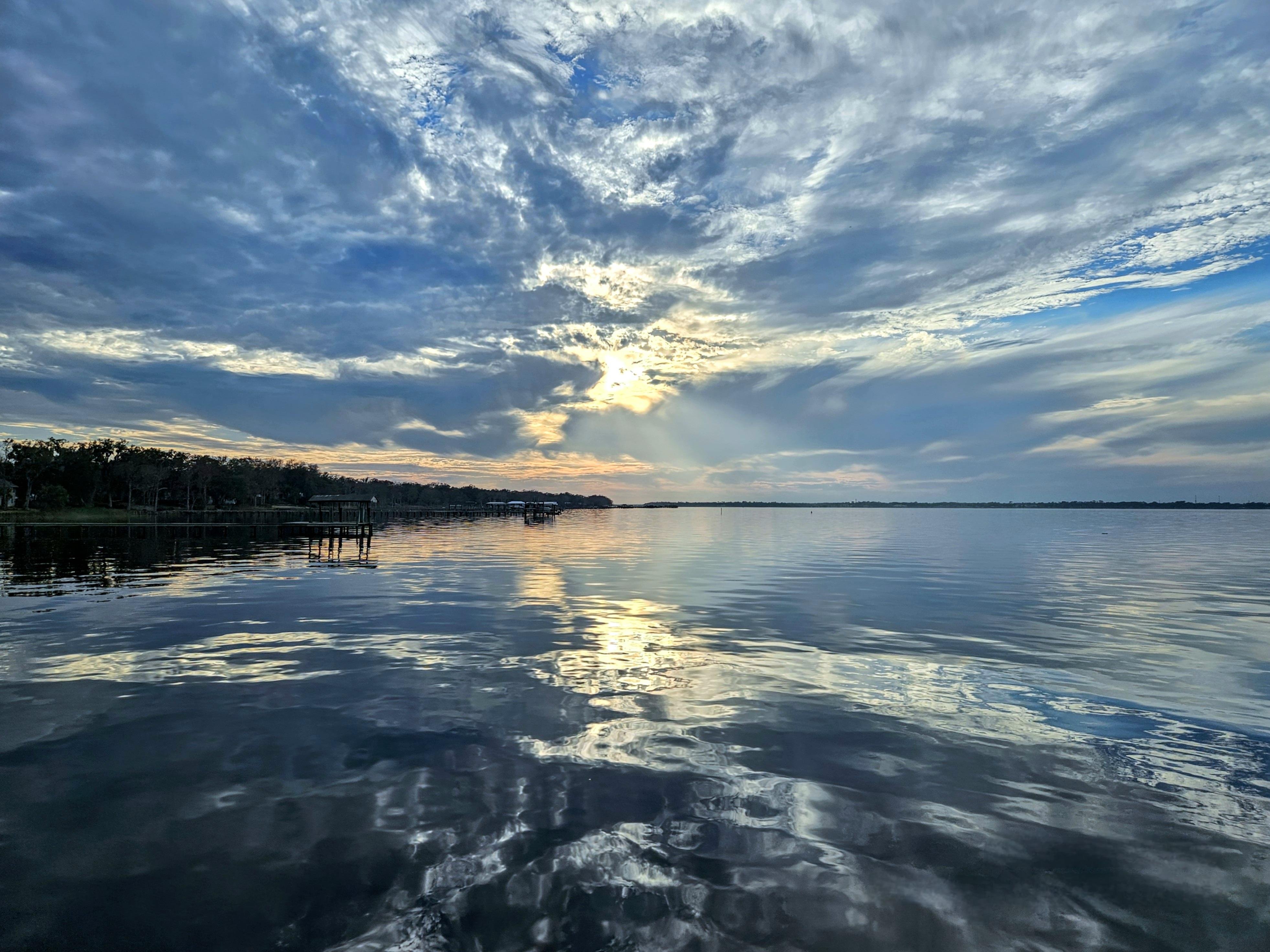 Calm river reflecting a dramatic, cloudy sky during sunset. Sunlight peeks through the clouds, creating a serene glow on the water. A tree-lined shore with docks is visible on the left, extending into the water.