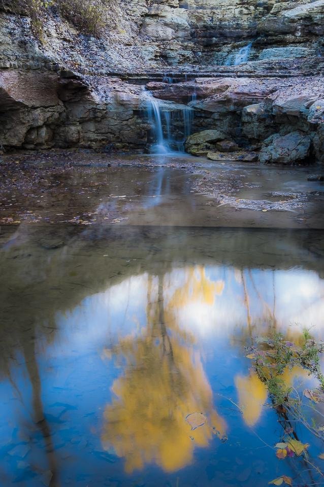 Small waterfall spilling over layered rock ledges into a shallow pool, with rocky canyon walls on both sides; calm water in the foreground reflects blue sky and yellow autumn tree branches, with a few leaves floating on the surface.