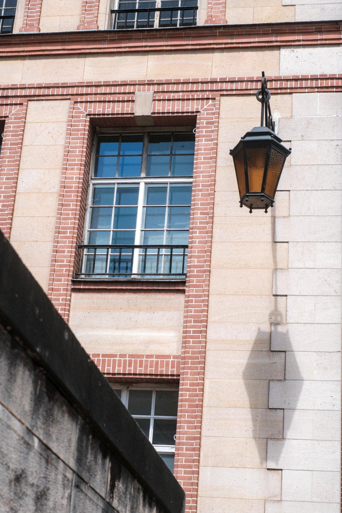 Photo verticale. Sur le mur d’un bâtiment dont le pourtour des fenêtres est décoré de petites briques rouges alors que le reste est beige, une lampe ornementale est fixée et son verre est orange. Le soleil projette son ombre sur le mur au 2 tons de beige. Sur la gauche, un bout de muret gris contraste la scène